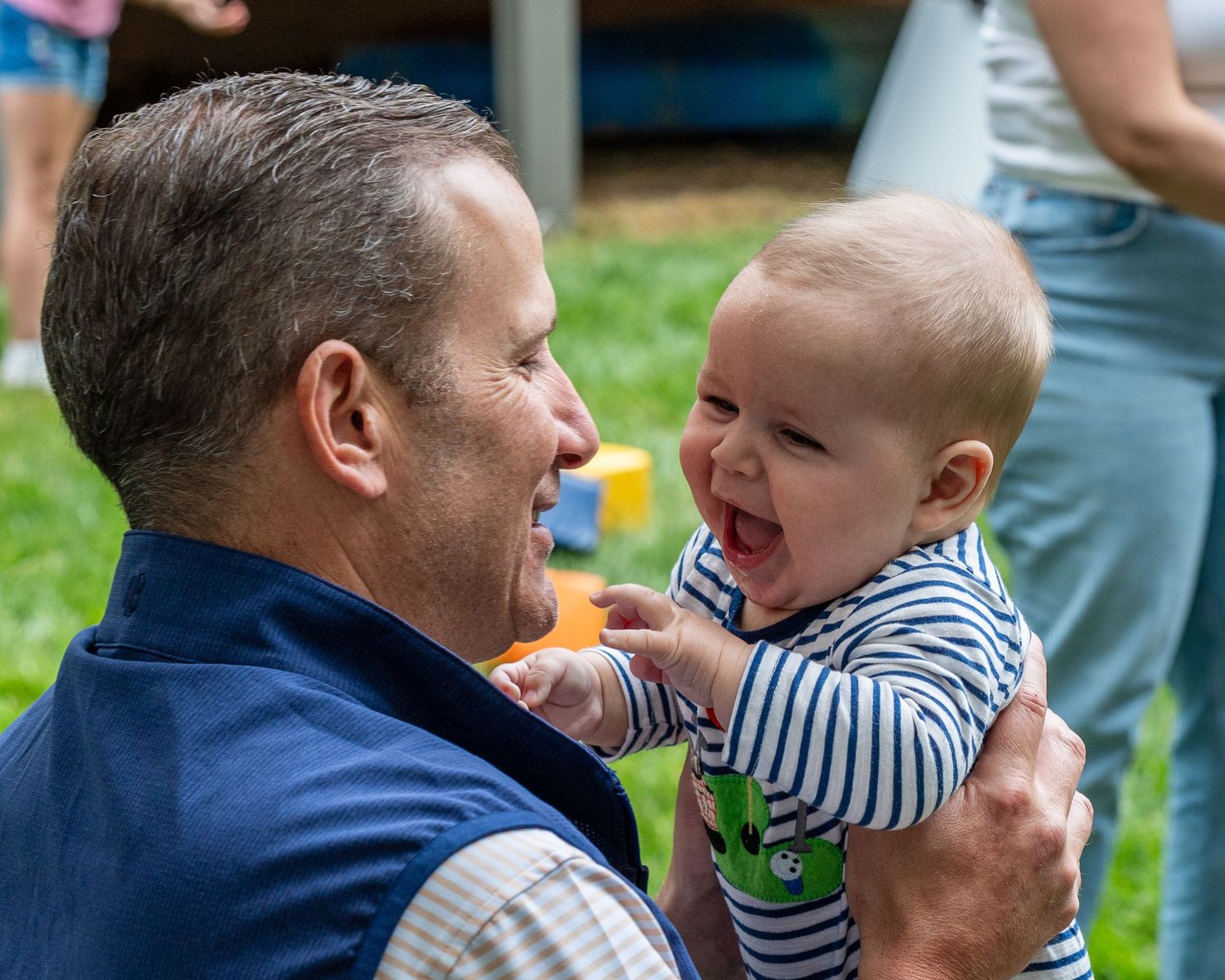 Smiling middle-aged man in blue vest holding a laughing baby outdoors at a sunny gathering.