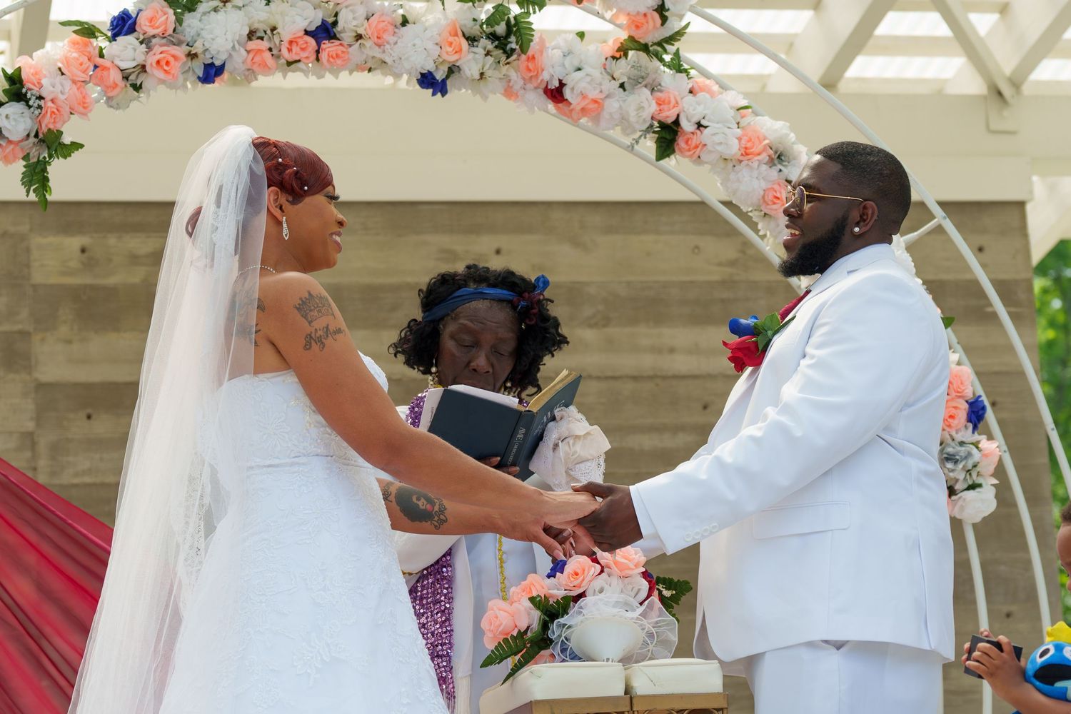 Black bride and groom holding hands at floral arch during outdoor wedding ceremony.
