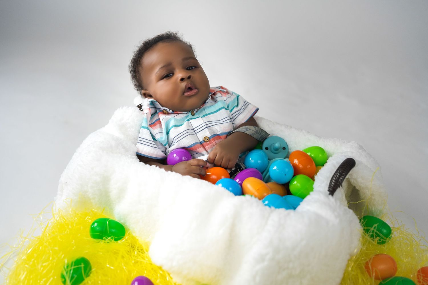 Adorable baby sitting in a fluffy Easter basket surrounded by colorful plastic eggs.