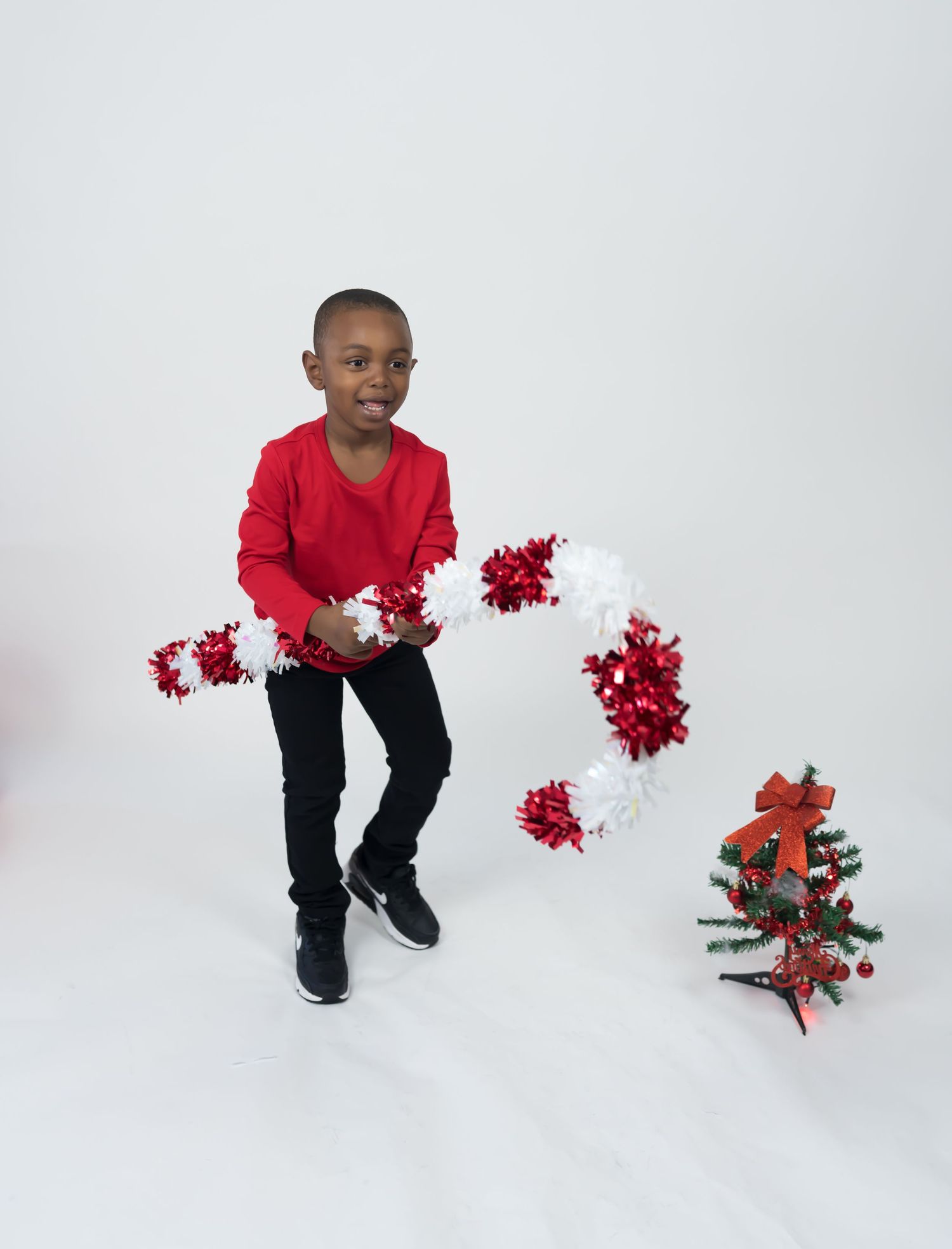 Young boy in red shirt holds red and white garland beside a small decorated Christmas tree.