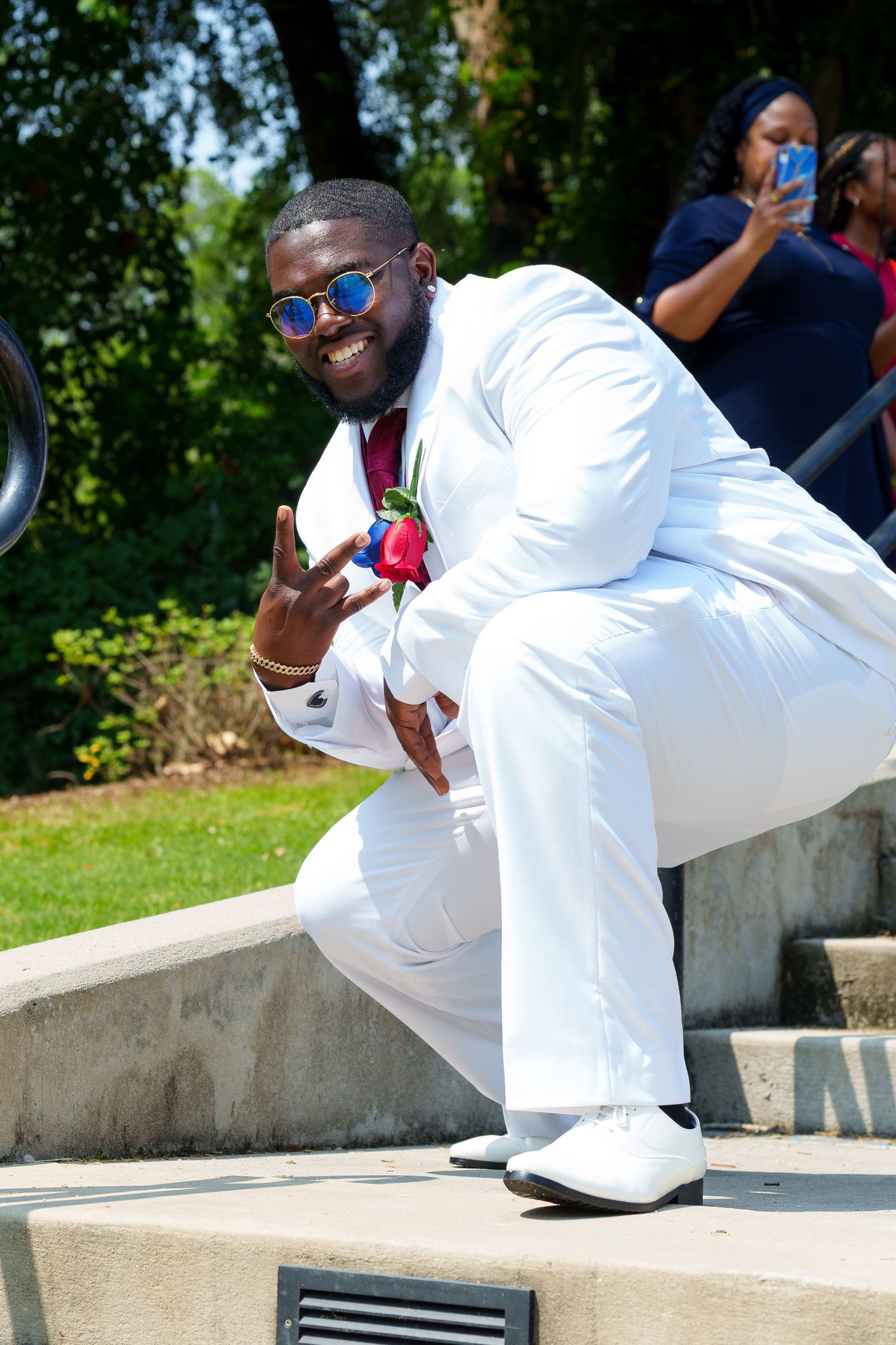 Smiling groom in white suit with floral boutonniere posing on steps at outdoor wedding venue.