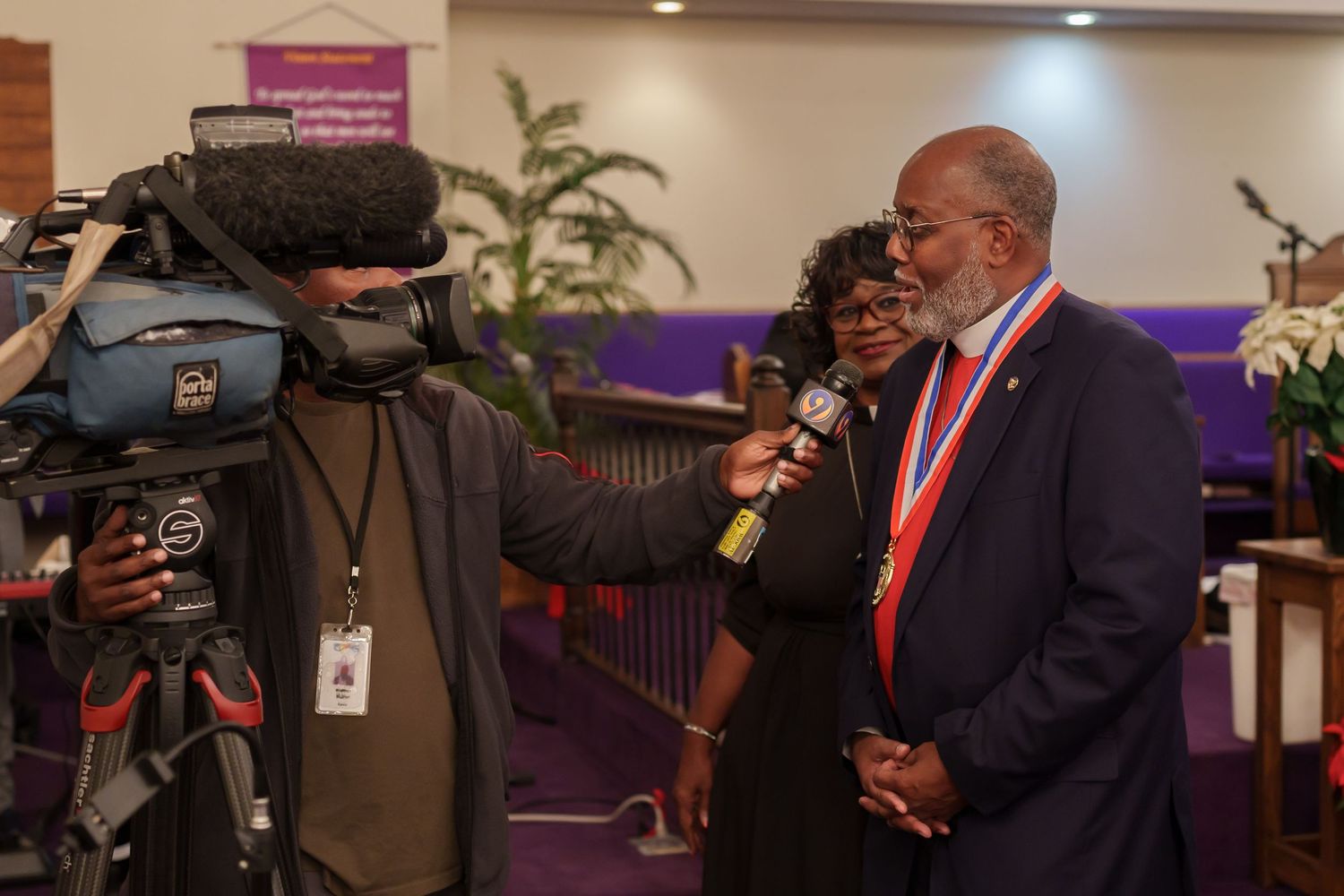 A man wearing a medal is interviewed by a TV camera operator inside a church.