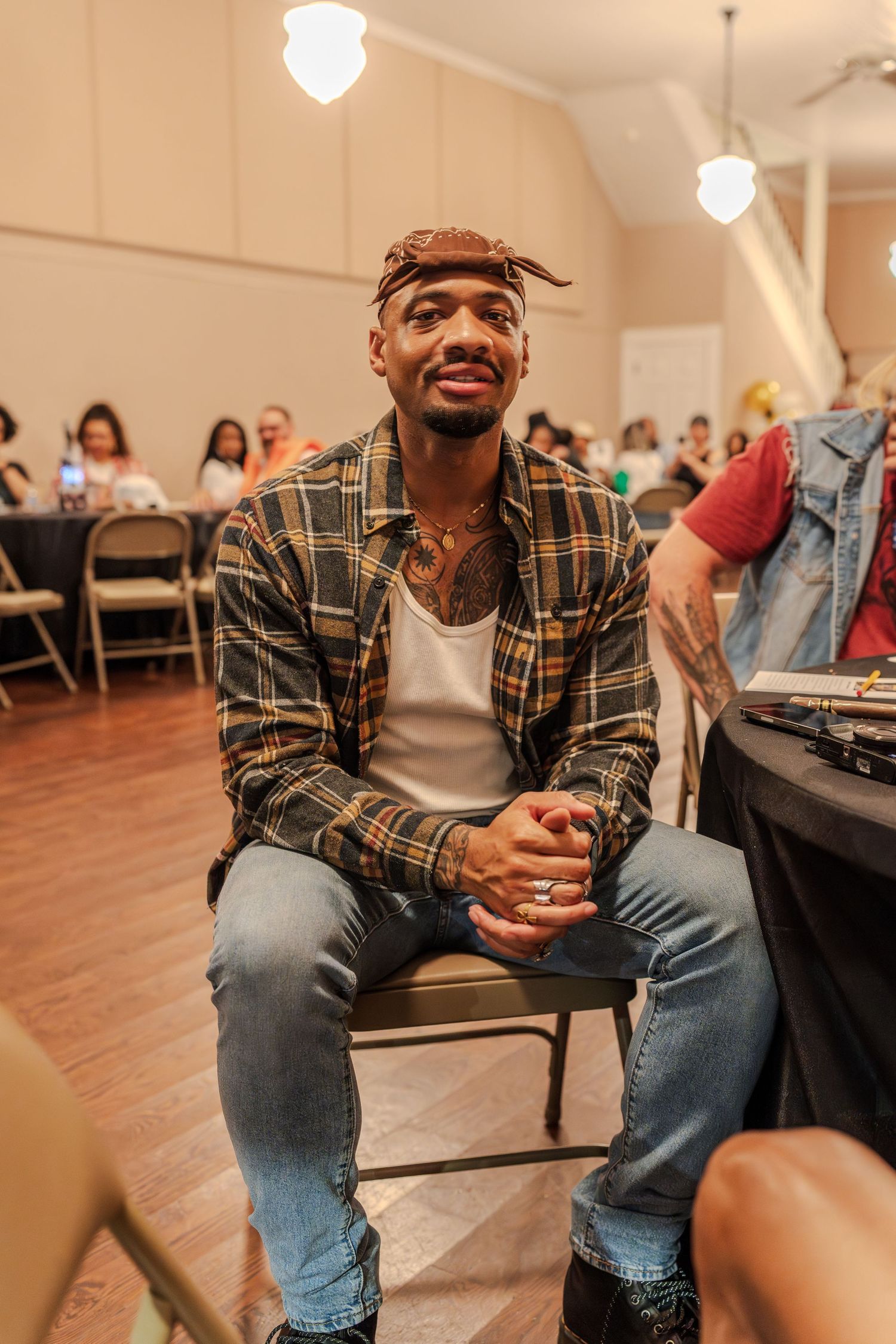 Smiling young Black man in plaid shirt and bandana sitting at a table during an indoor event.
