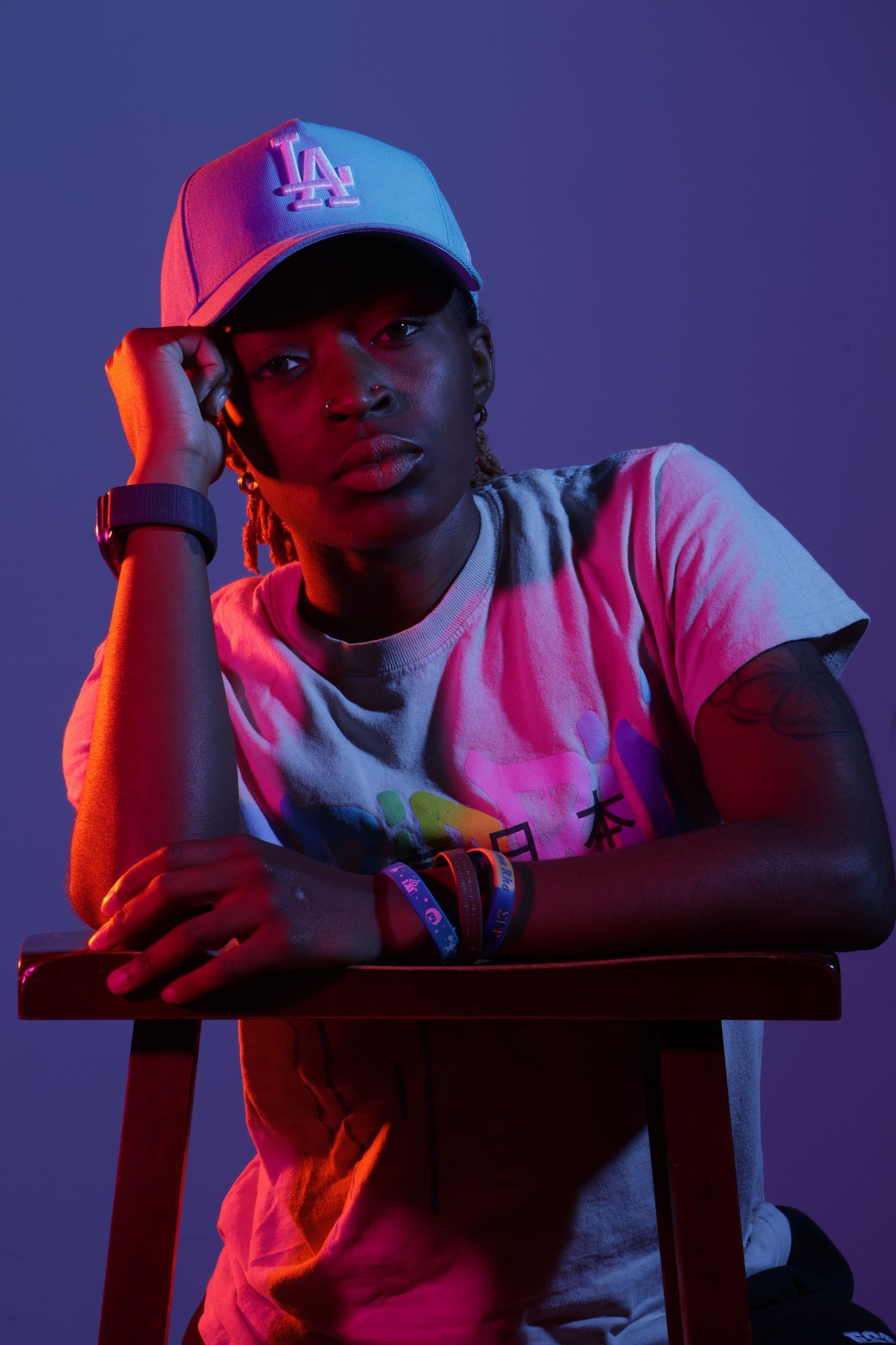 A young man in an LA Dodgers cap poses on a chair under dramatic neon purple and pink studio lighting.