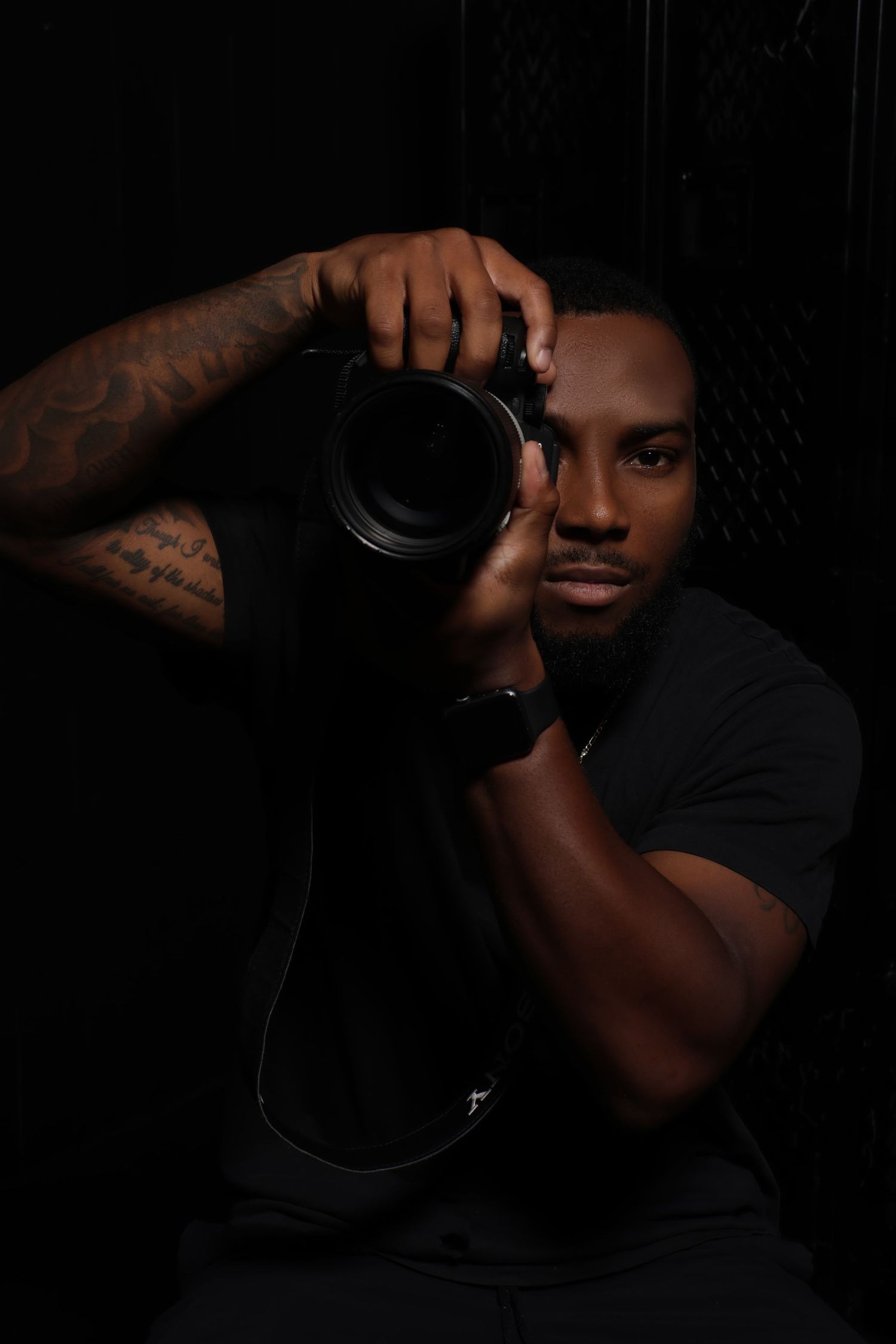 Male photographer with tattoos aiming DSLR camera toward viewer in dramatic dark studio portrait.