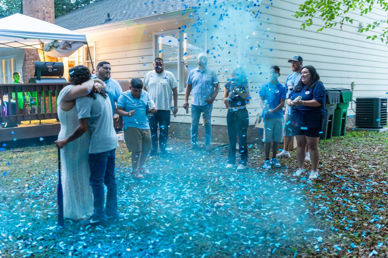 Couple embracing in blue confetti cloud at outdoor gender reveal party surrounded by cheering family.