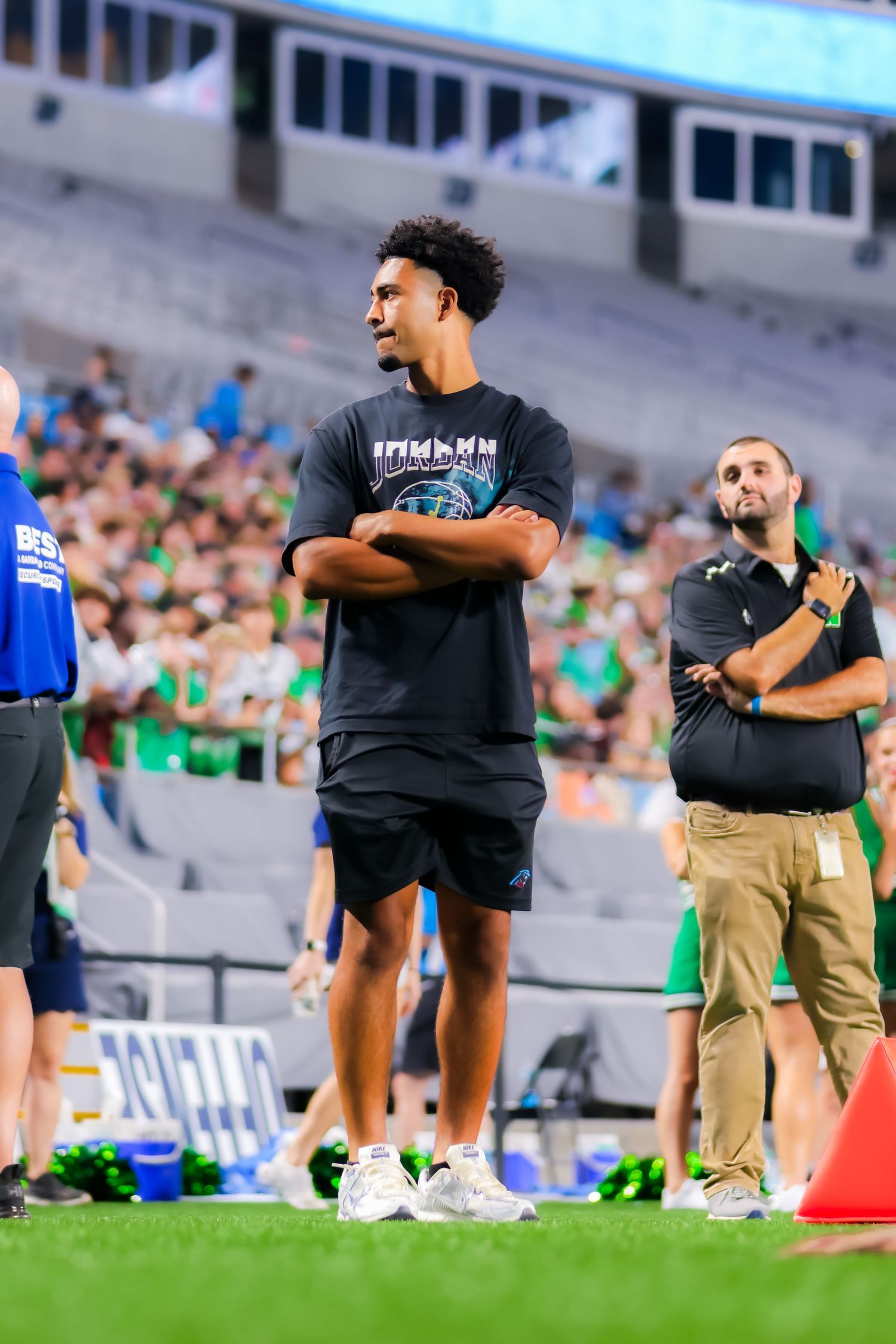 Tall young man with crossed arms stands on sideline watching college football game in packed stadium.