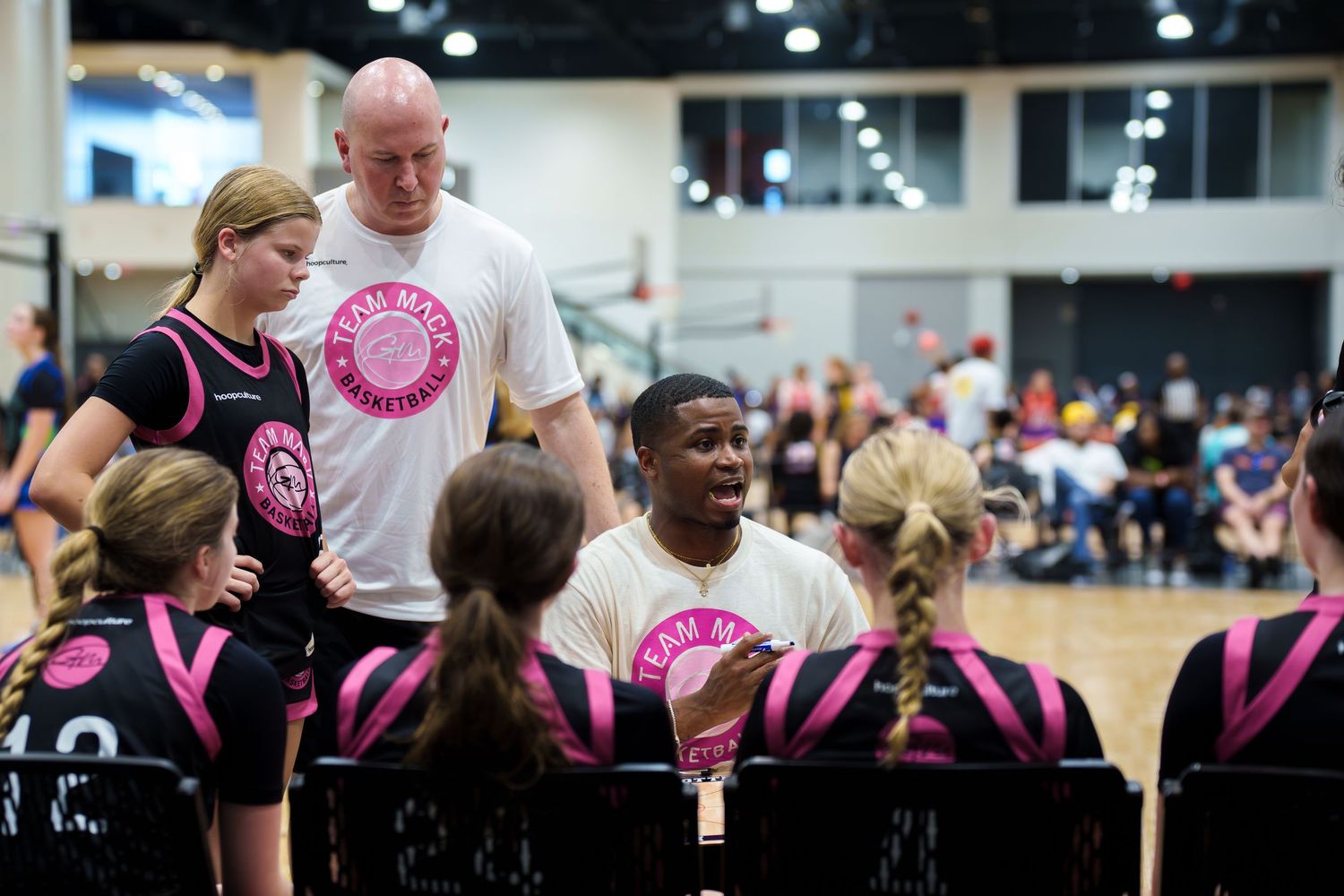 Coach in white shirt talking to young girls in pink uniforms during a basketball timeout.