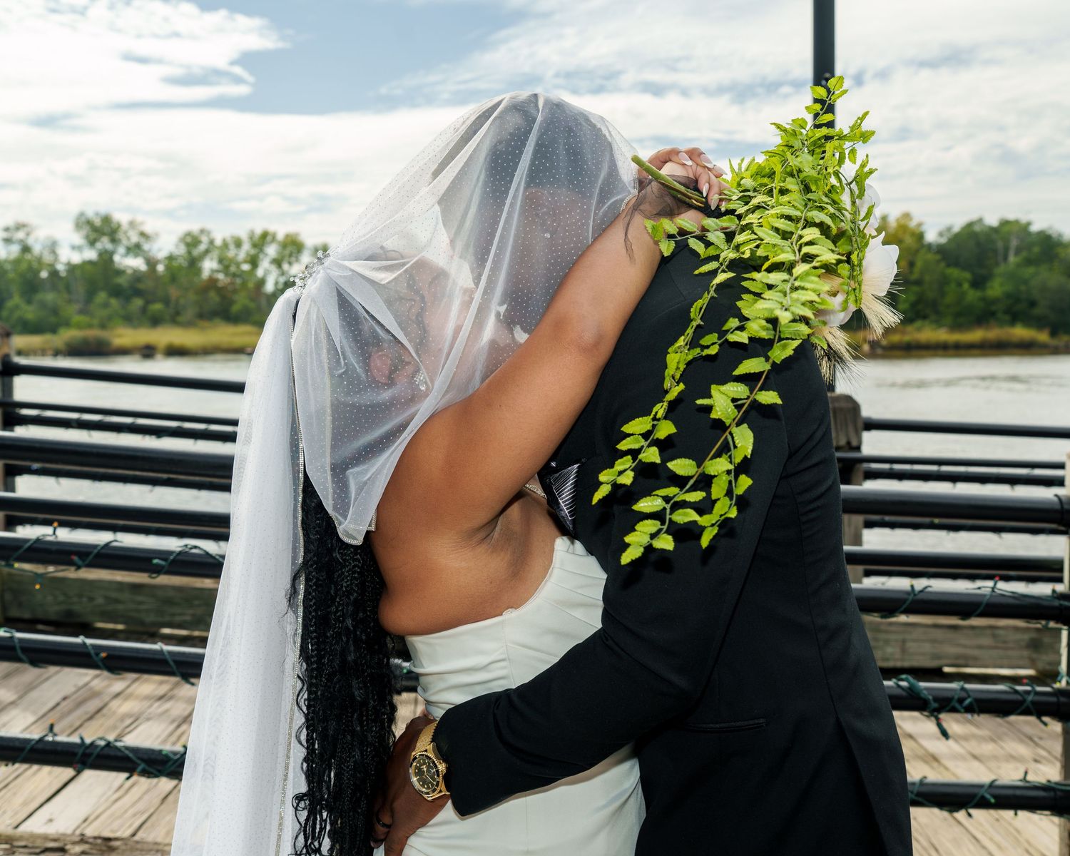 Bride and groom share a romantic kiss on a waterfront dock, bride wearing a flowing white veil and gown.