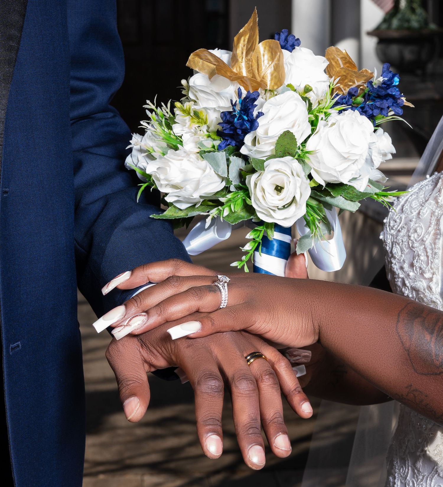 Newlywed couple holding white and blue wedding bouquet with gold accents, showing wedding rings on their hands.