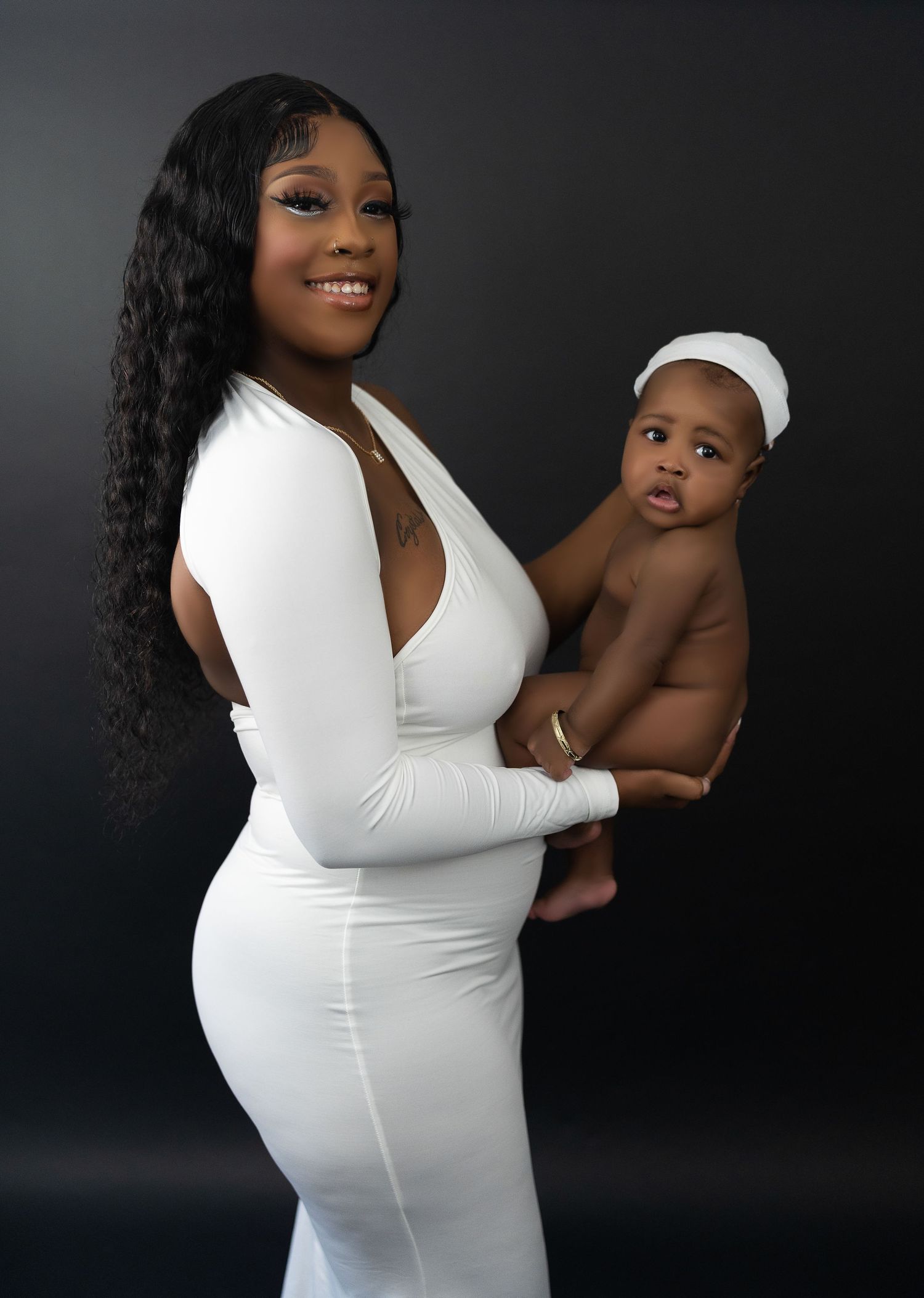 Smiling mother in white dress holds baby with white headband in dark studio portrait.