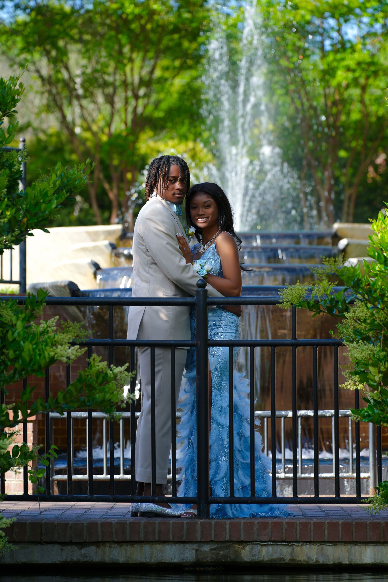 Prom couple in tan suit and blue sequin gown embracing near fountain in garden park.