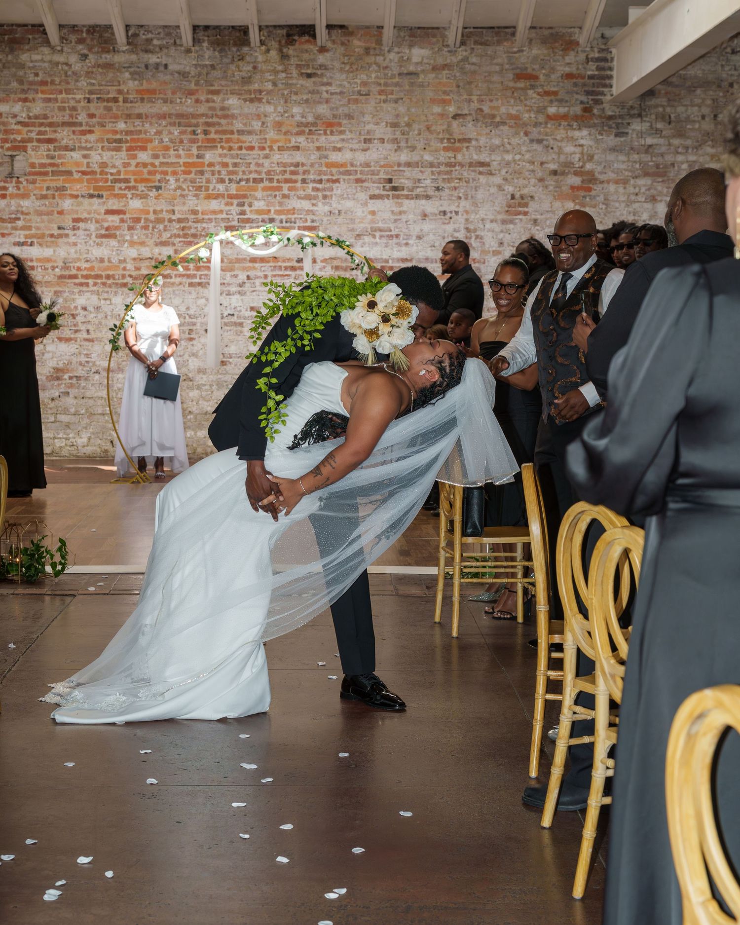 Groom dips bride in white gown and veil during wedding ceremony kiss before cheering guests.