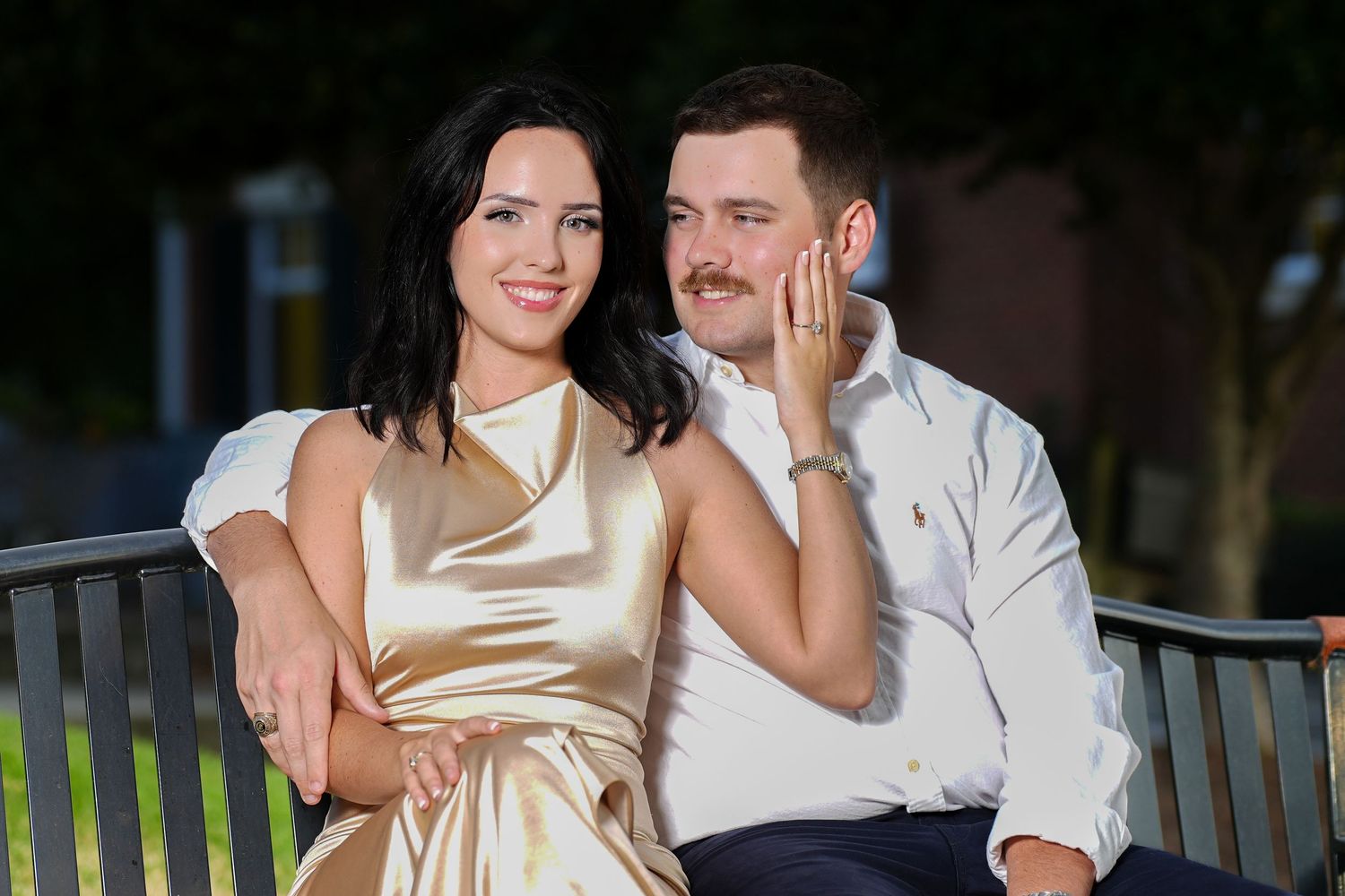Couple posing on a bench outdoors, woman in gold dress and man in white shirt smiling together.