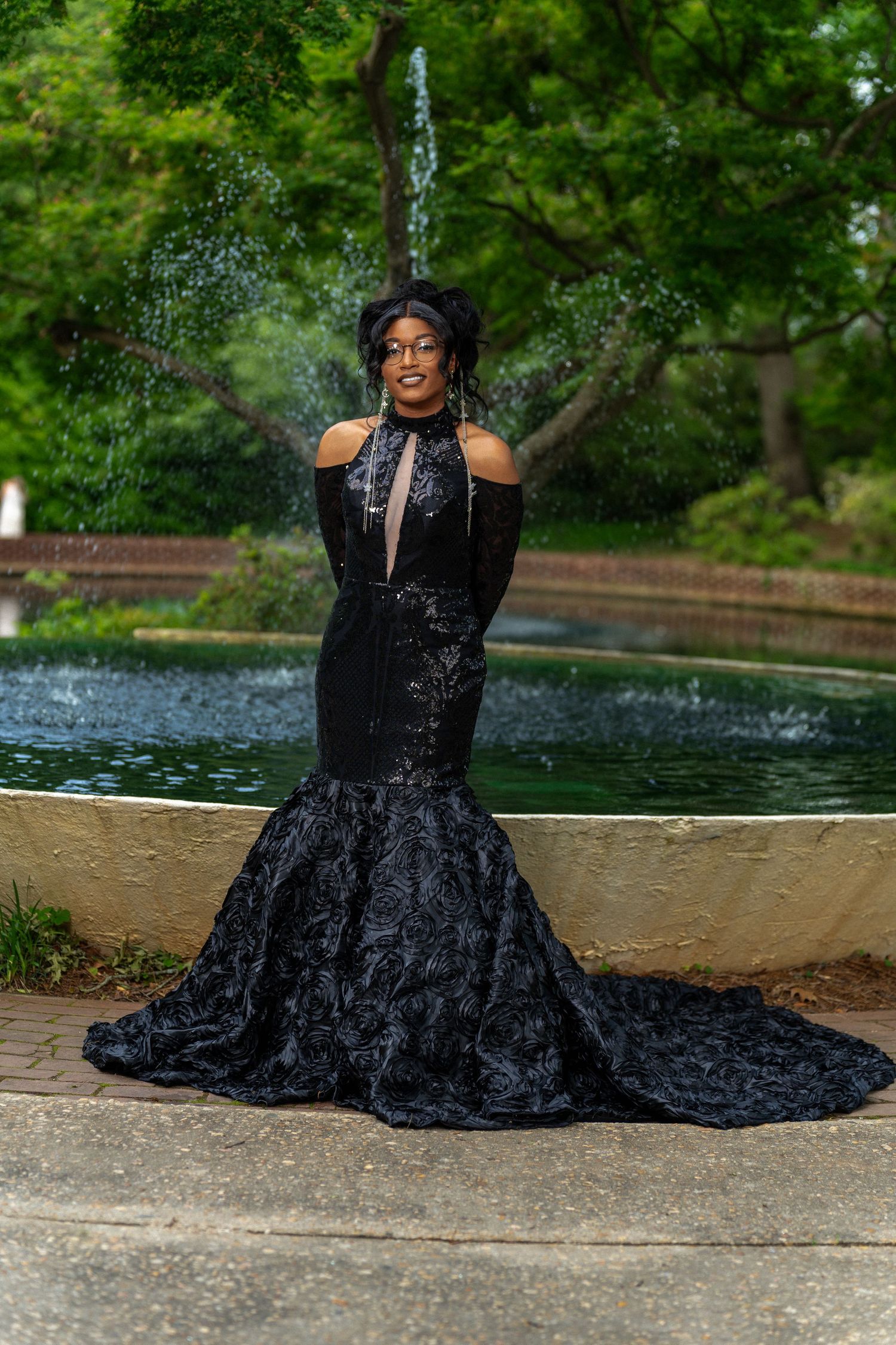 Young woman in stunning black mermaid gown with train posing by garden fountain at prom.