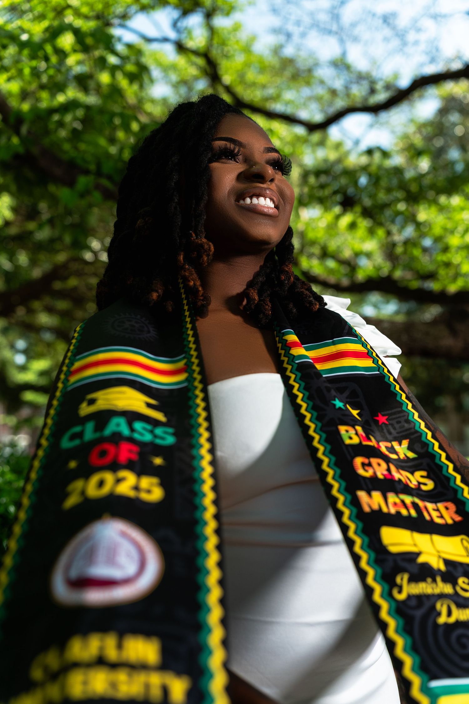 Smiling Black graduate wearing a Claflin University Class of 2025 kente stole and Black Grads Matter sash outdoors.