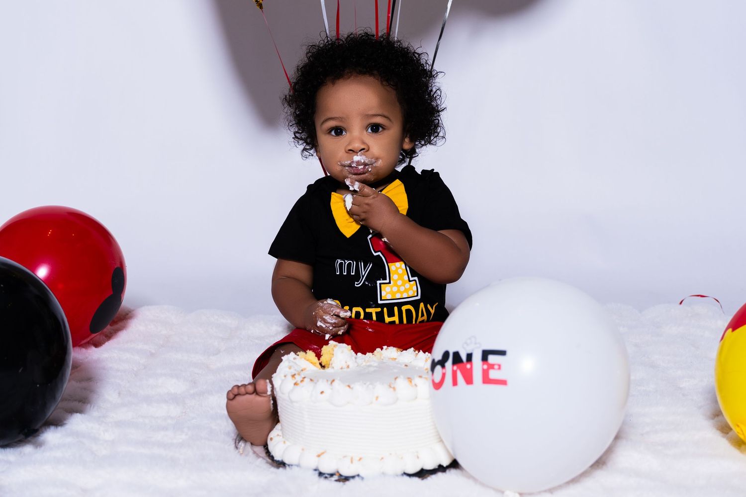 Baby boy smashing first birthday cake surrounded by red black and white balloons.