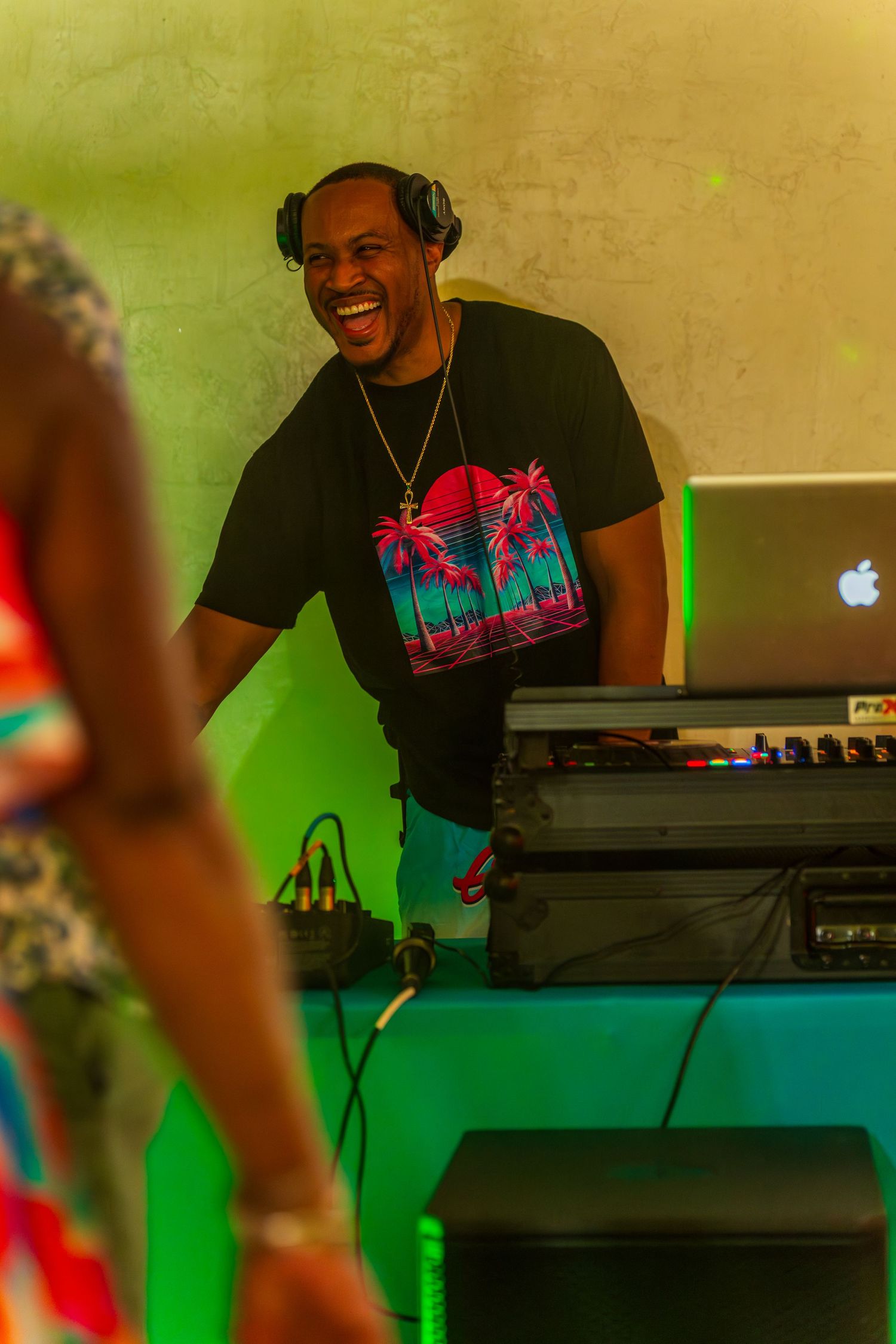 Smiling DJ wearing headphones and tropical t-shirt mixing music on laptop at colorful neon-lit party event.