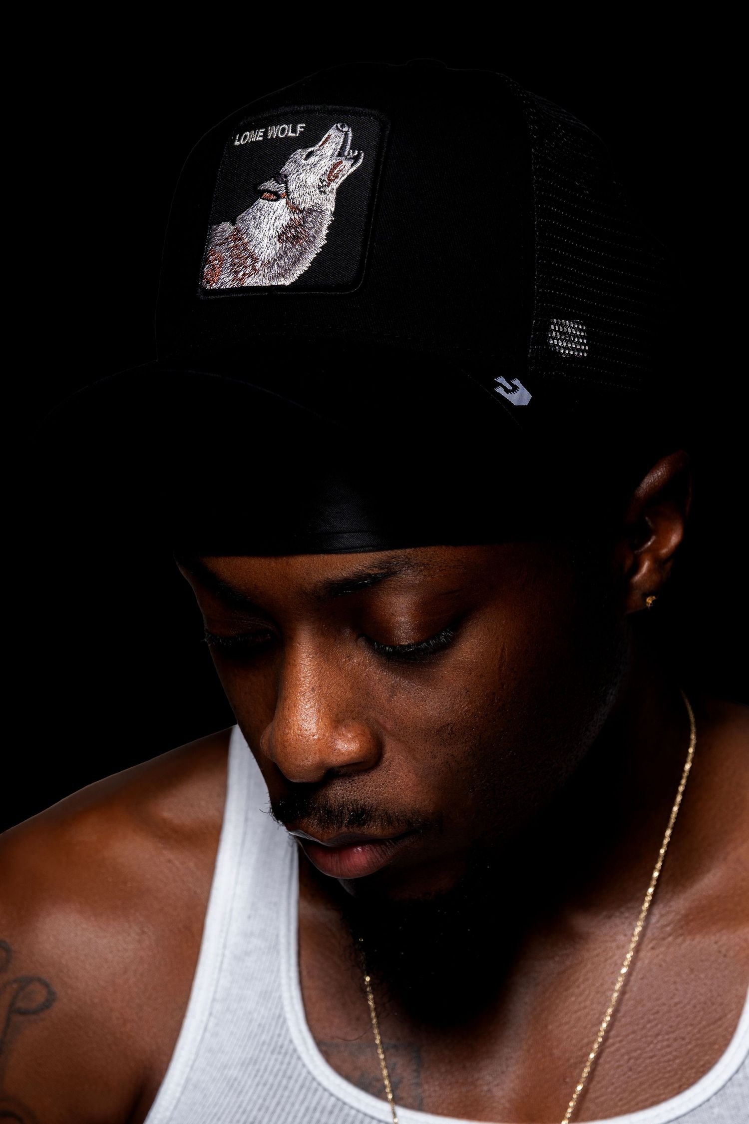 Young man in white tank top wearing black Lone Wolf trucker hat looking down.