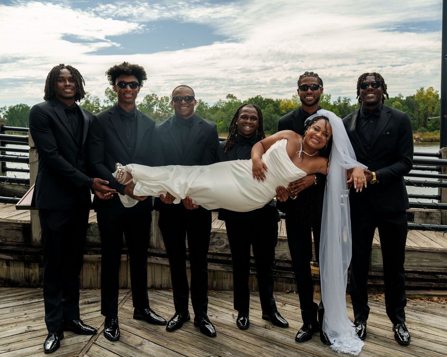 Groom and groomsmen in black suits lifting smiling bride in white gown on outdoor wooden dock.