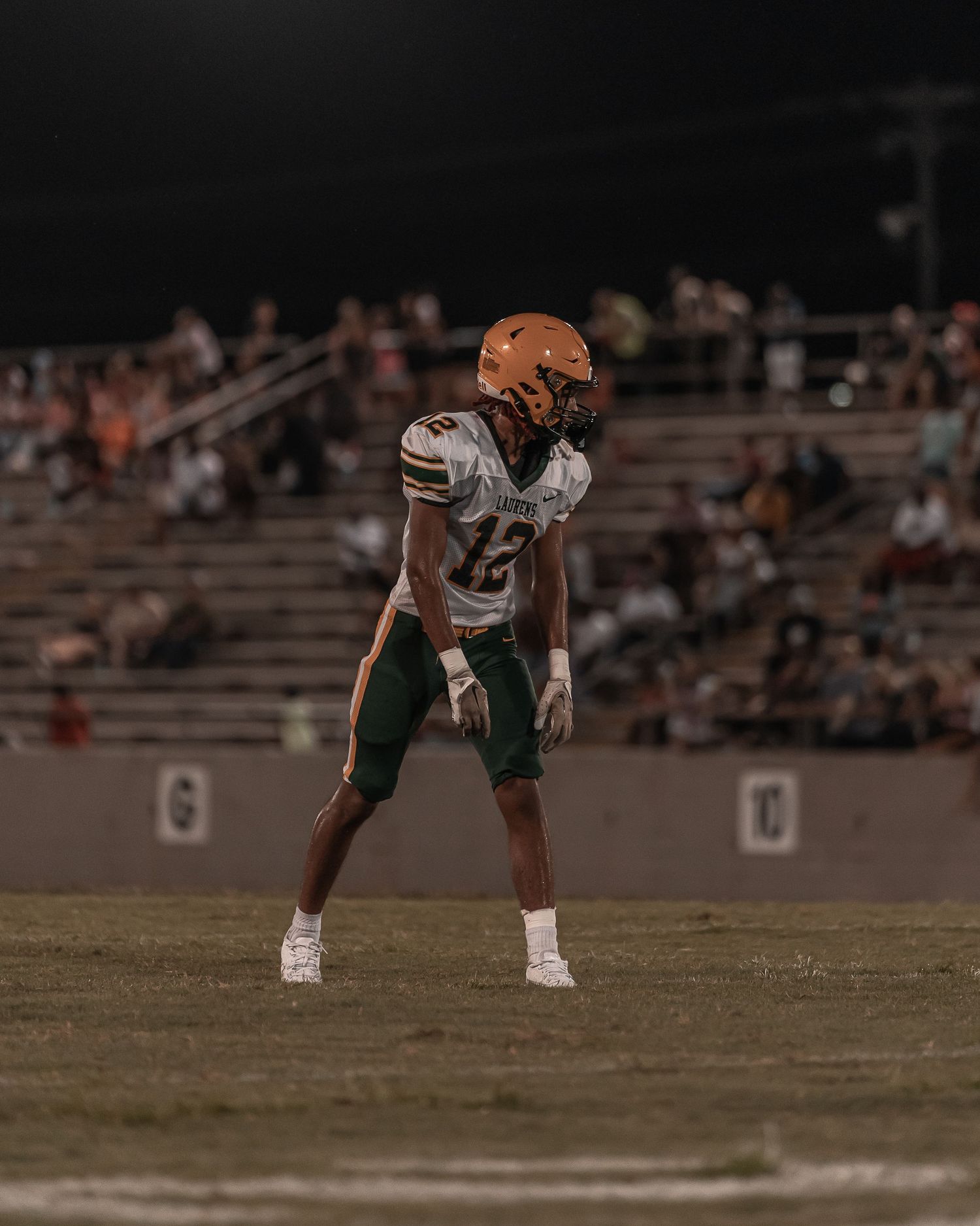 High school football player number 12 in green and orange uniform in defensive stance at night game.