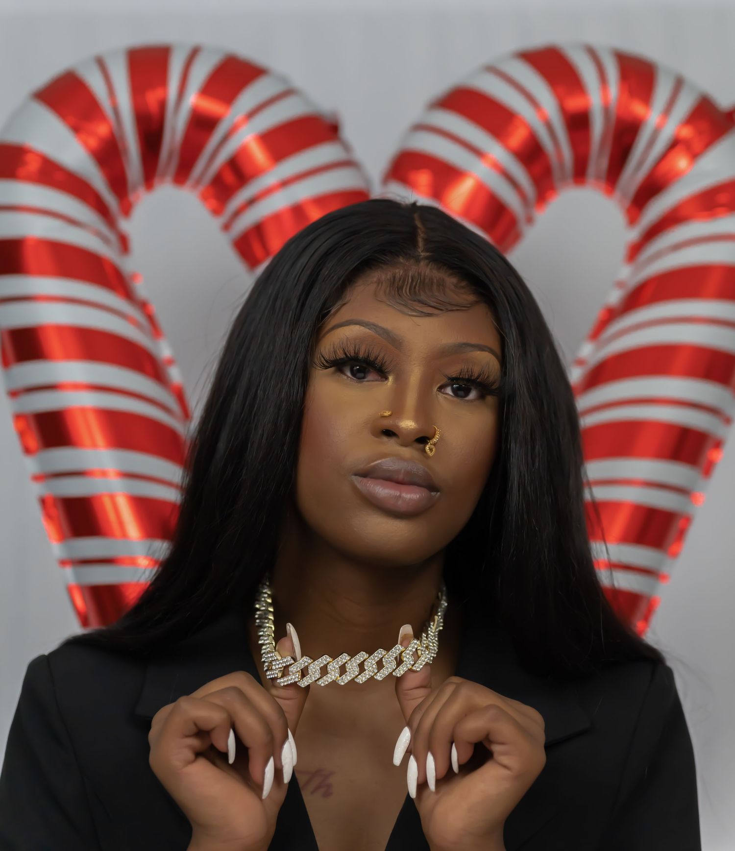 Woman wearing a gold Cuban link choker poses in front of red candy cane heart balloons.