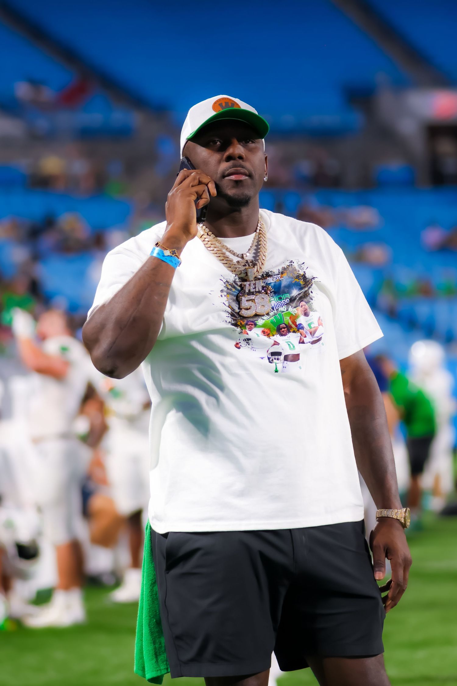 Man in white graphic tee and gold chain talks on phone at football stadium.