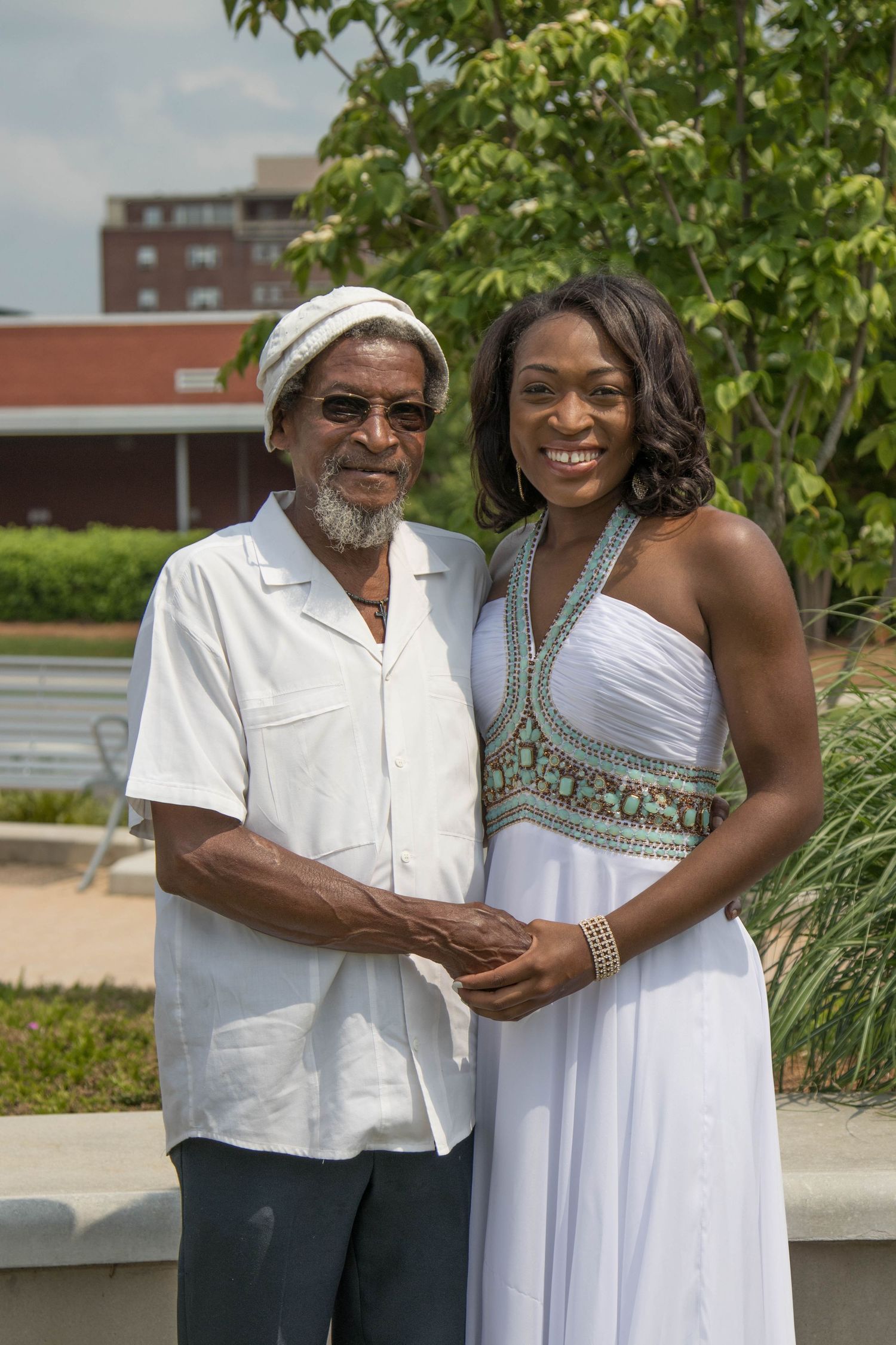 Elderly man and woman in white outfits smiling together outdoors near green trees.