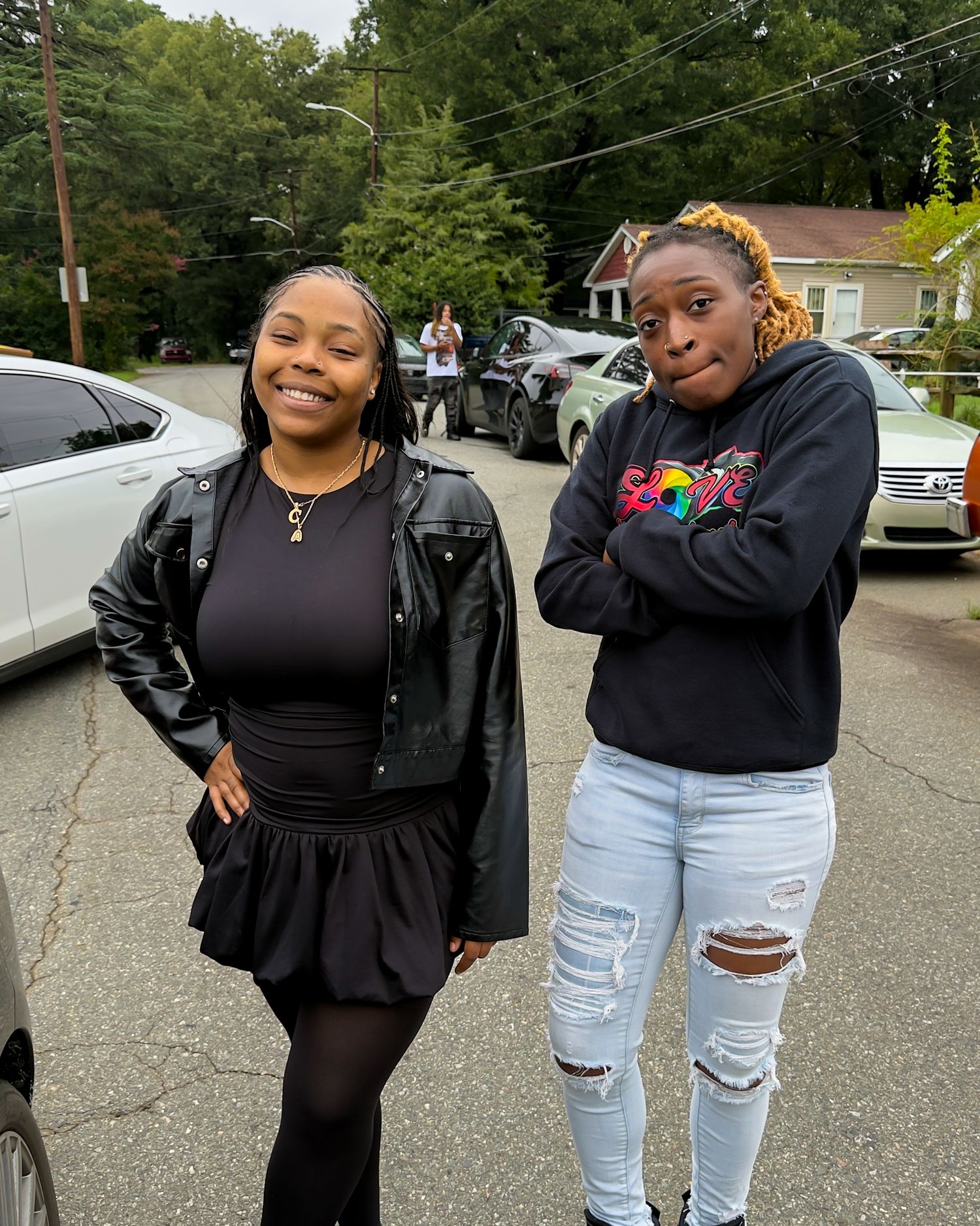 Two young women posing outdoors on a residential street near parked cars.