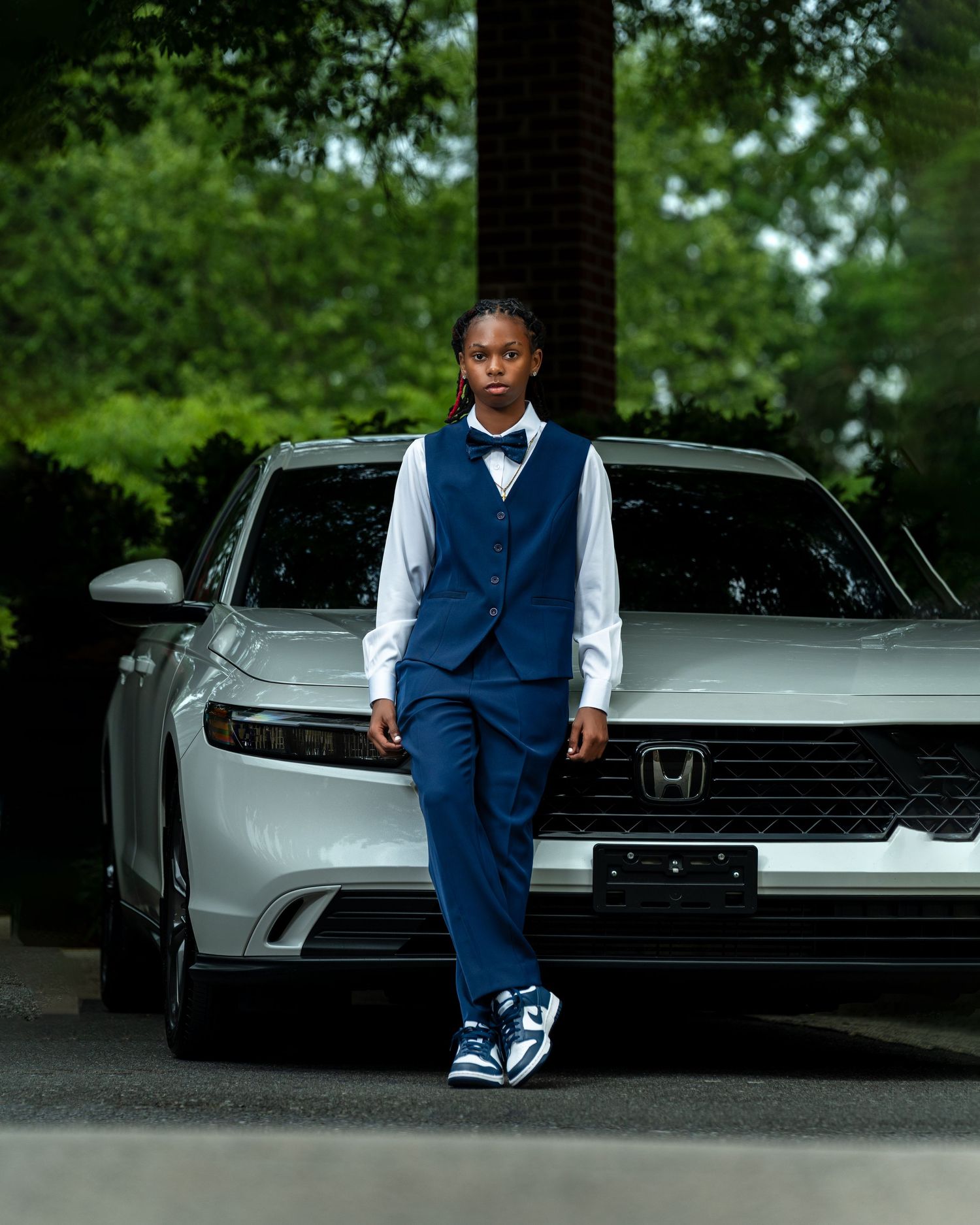 Young person in navy blue suit with bow tie leaning against silver Honda car outdoors.