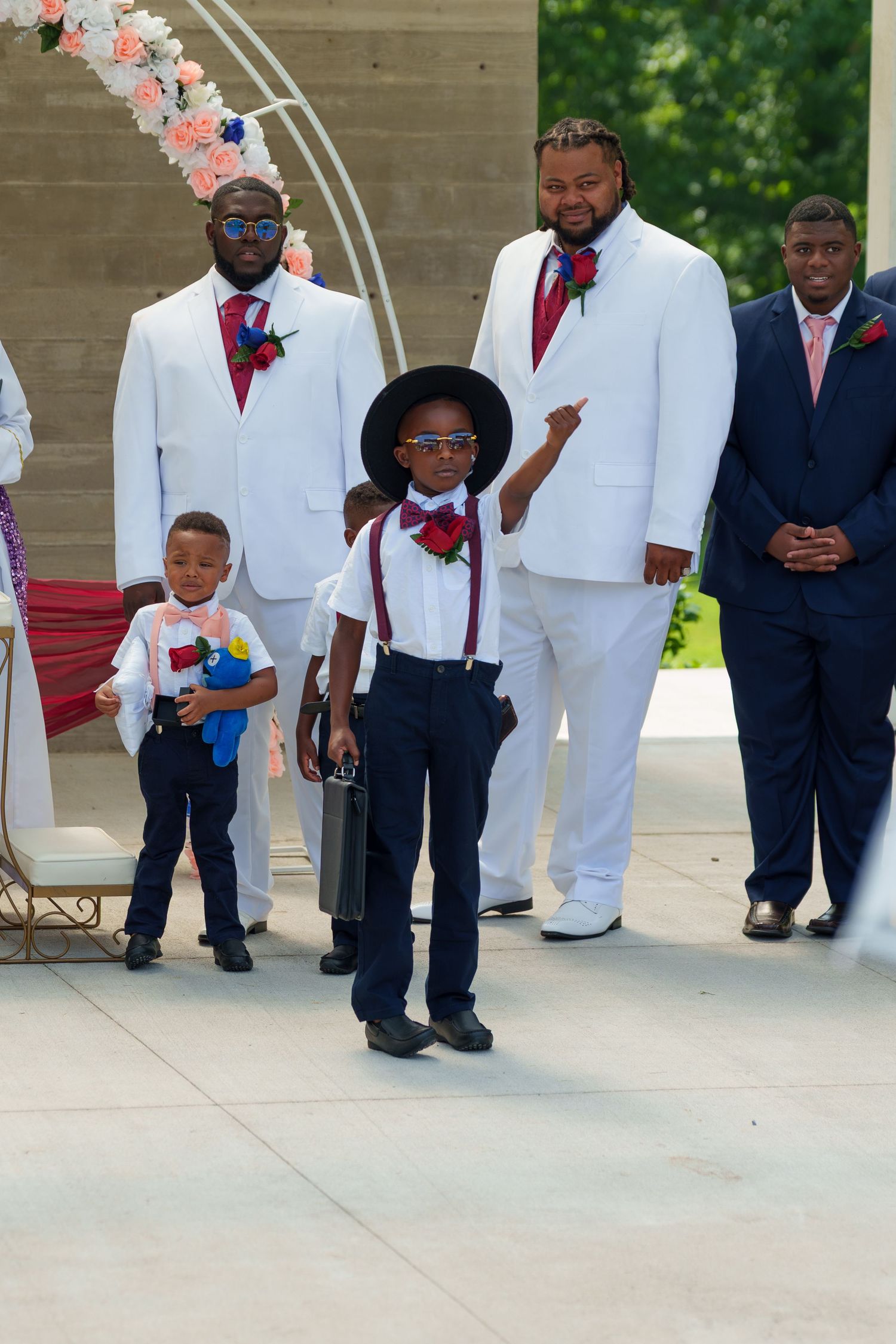 Ring bearer in hat and suspenders pointing at camera with groomsmen in white suits behind.