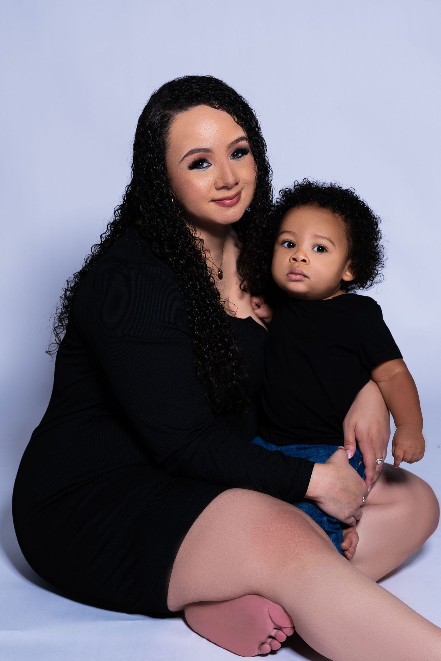 Mother with long curly hair sits with toddler in matching black outfits in studio portrait.