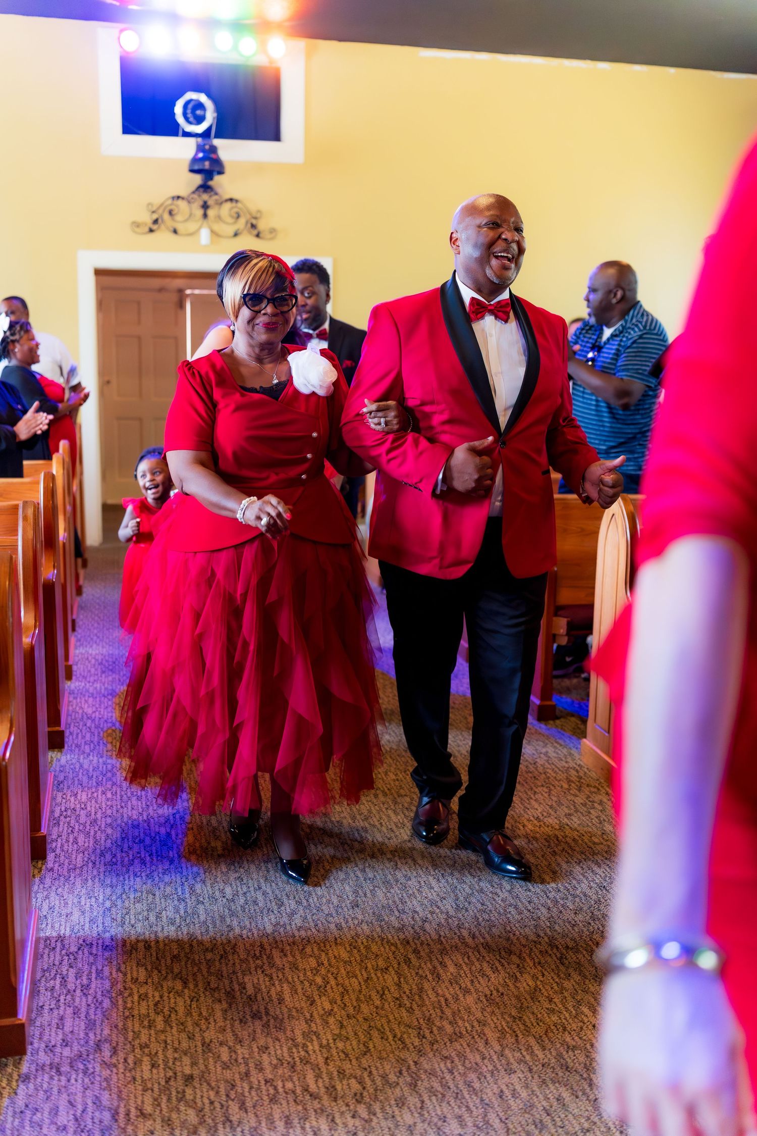 Couple in matching red outfits dancing joyfully down church aisle during celebratory ceremony.