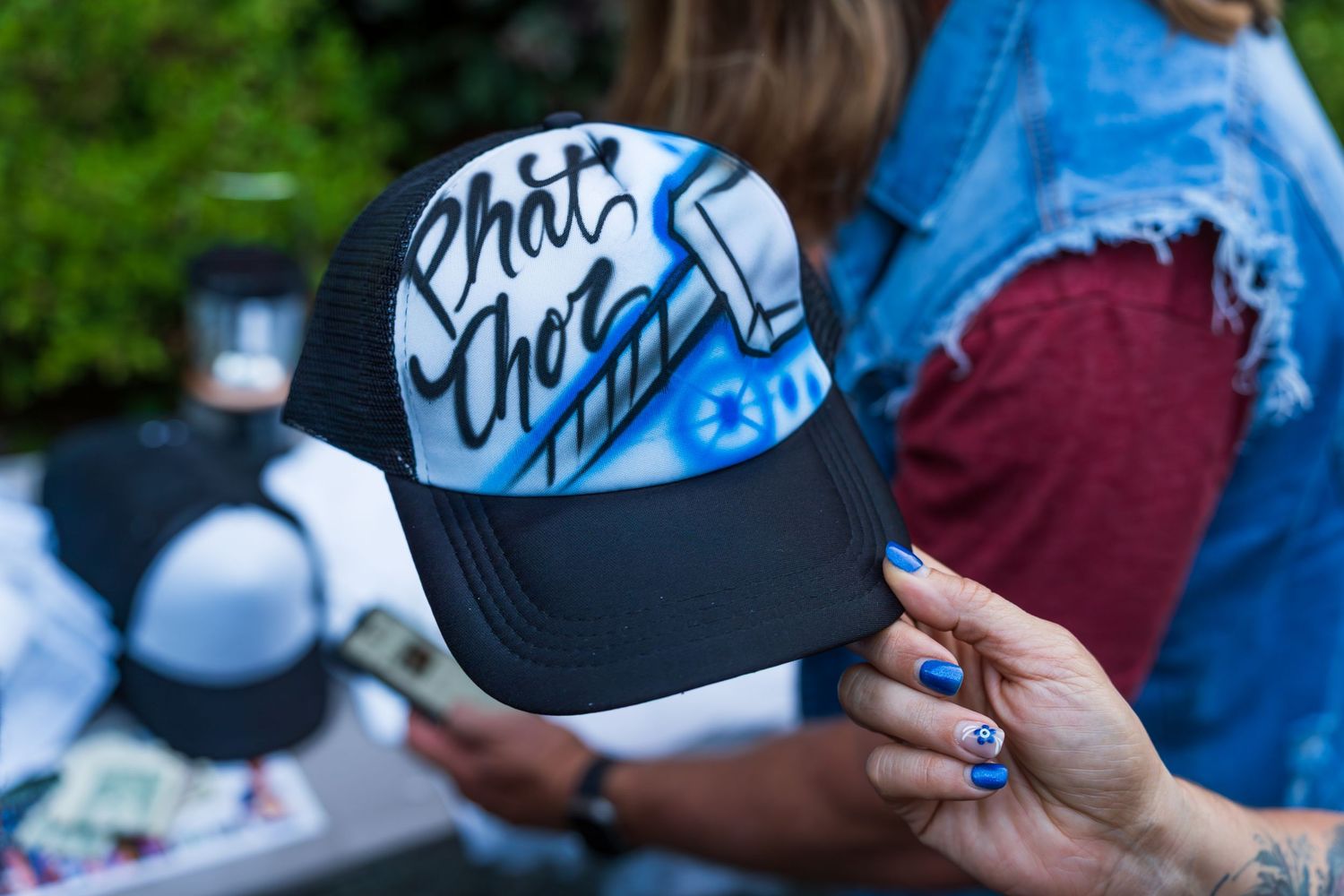 Hand with blue nails holding a black trucker hat with white and blue airbrush graffiti design outdoors.