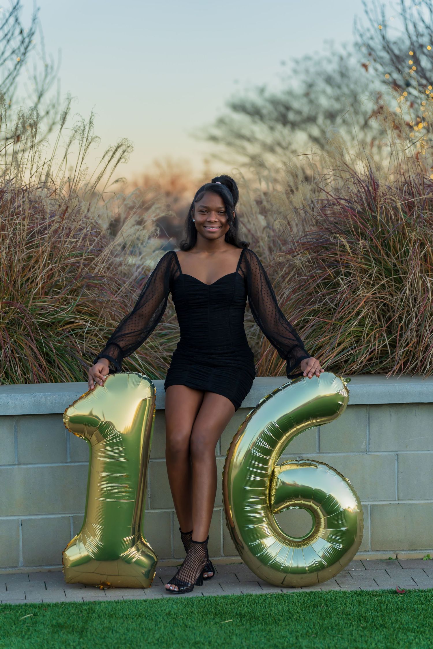 Smiling teen girl in black dress posing with large gold number 16 balloons at golden hour.