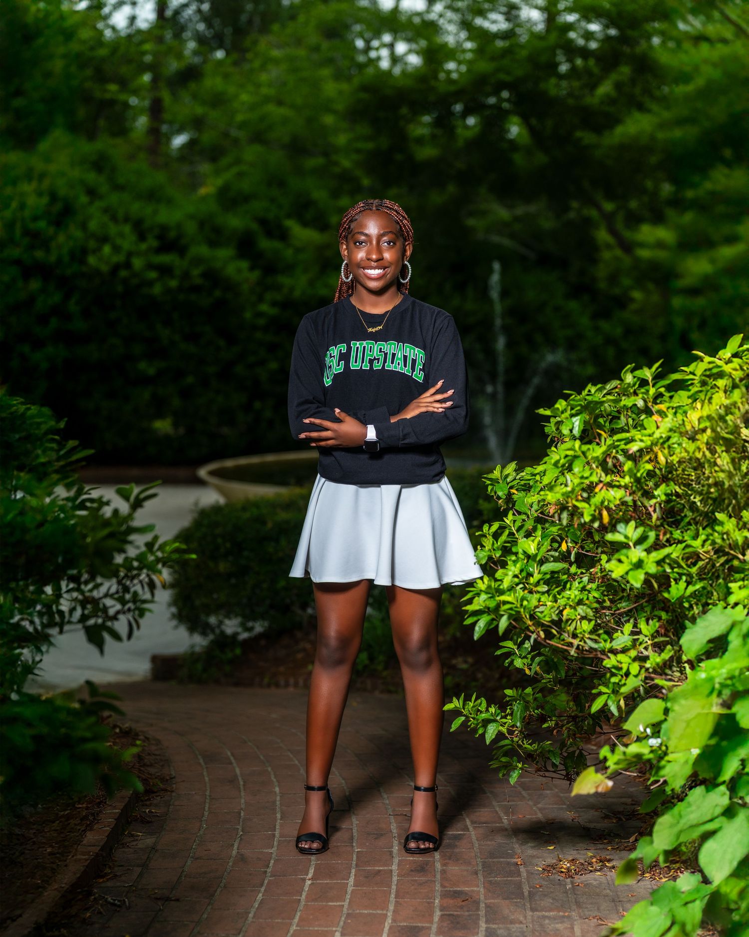 Confident young Black woman in SC Upstate sweatshirt and white skirt posing arms crossed in garden setting.