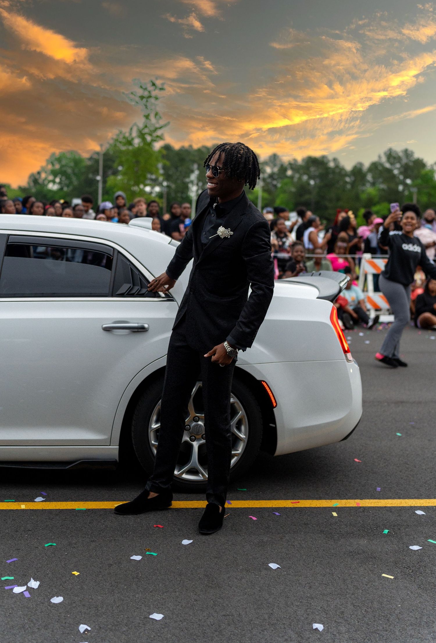 Young man in black suit leaning against white Cadillac at prom grand march with crowd at sunset.