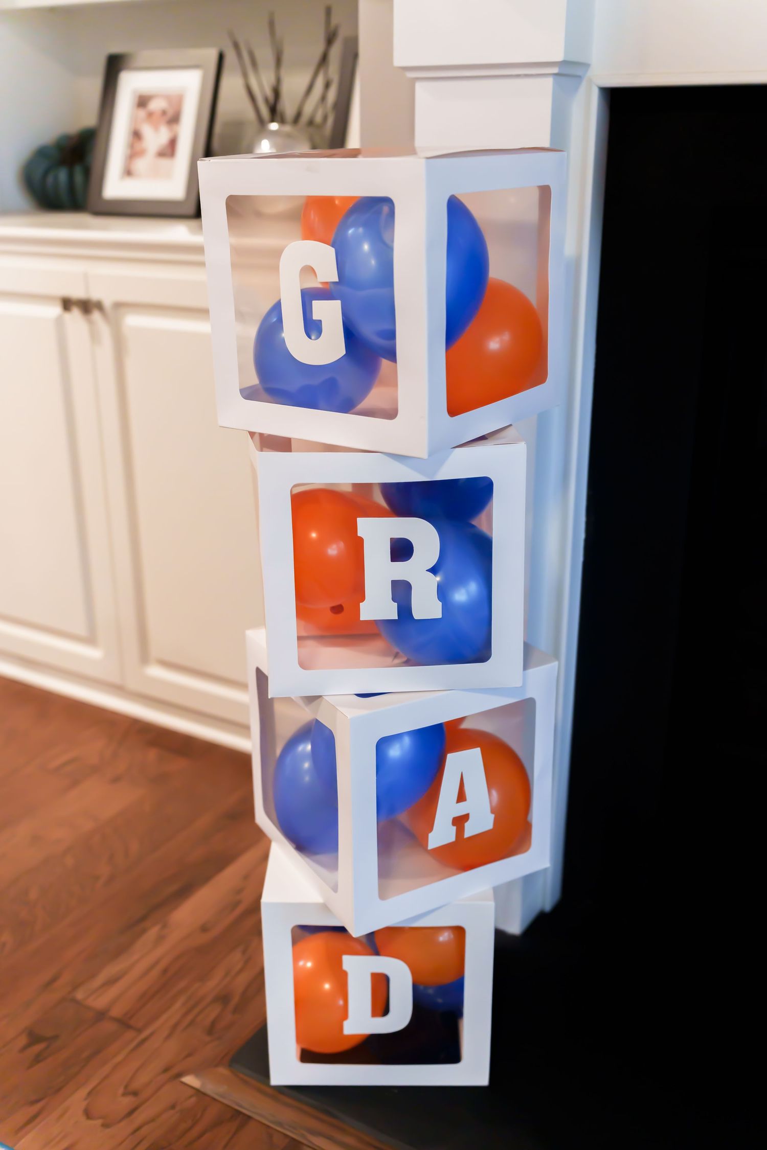 White transparent boxes stacked spelling GRAD filled with blue and orange balloons for graduation.