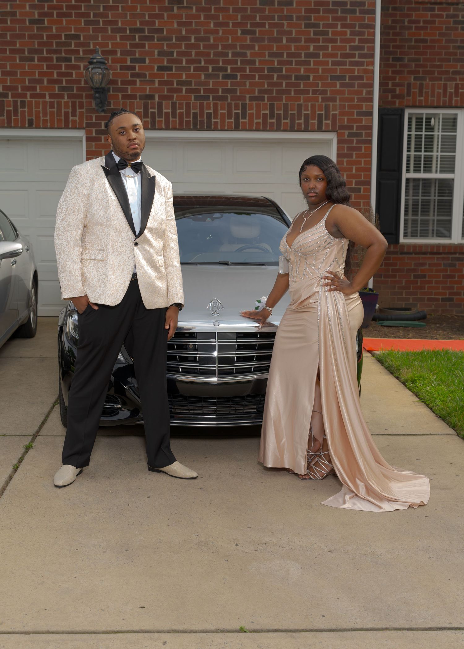 Prom couple in formal attire posing in front of silver Mercedes-Benz in residential driveway.
