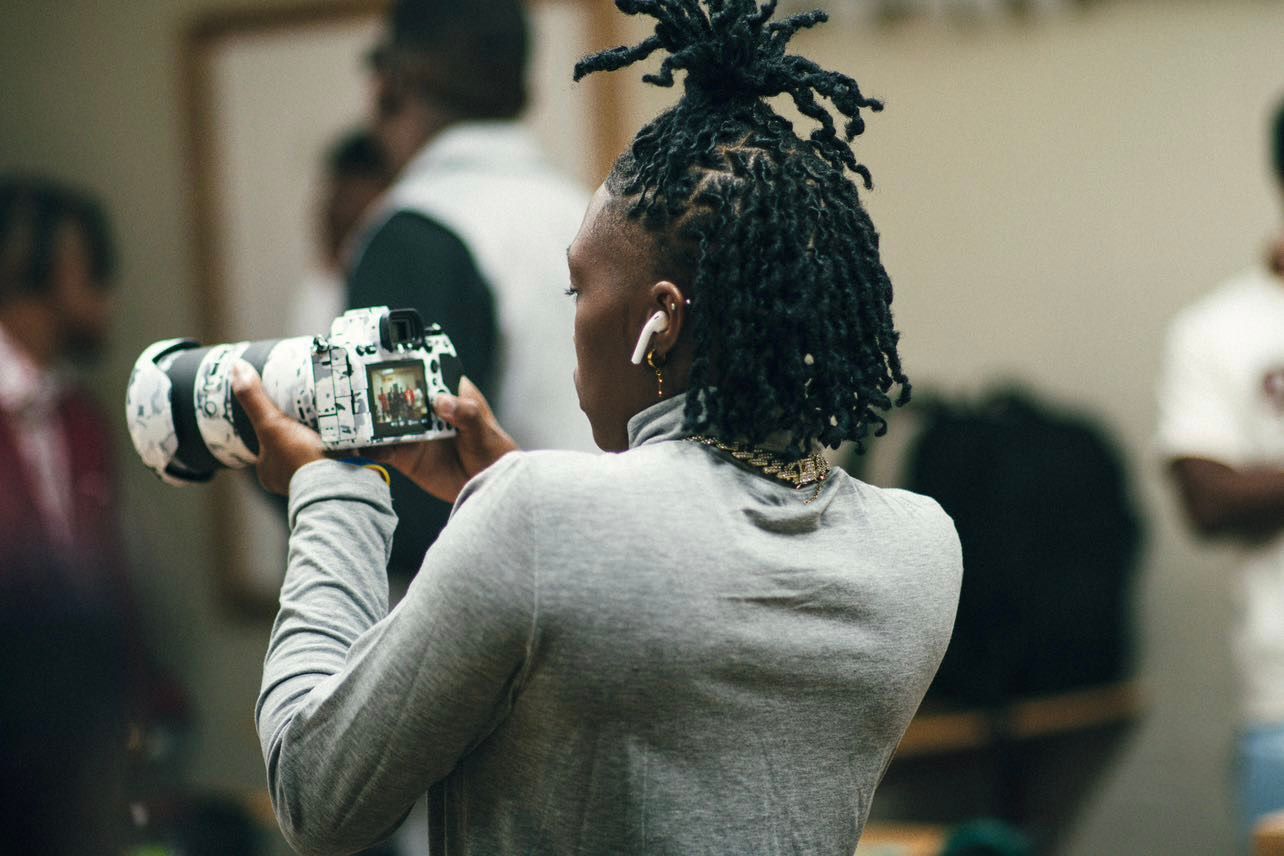 Young person with locs uses smartphone to photograph a supplement container indoors.