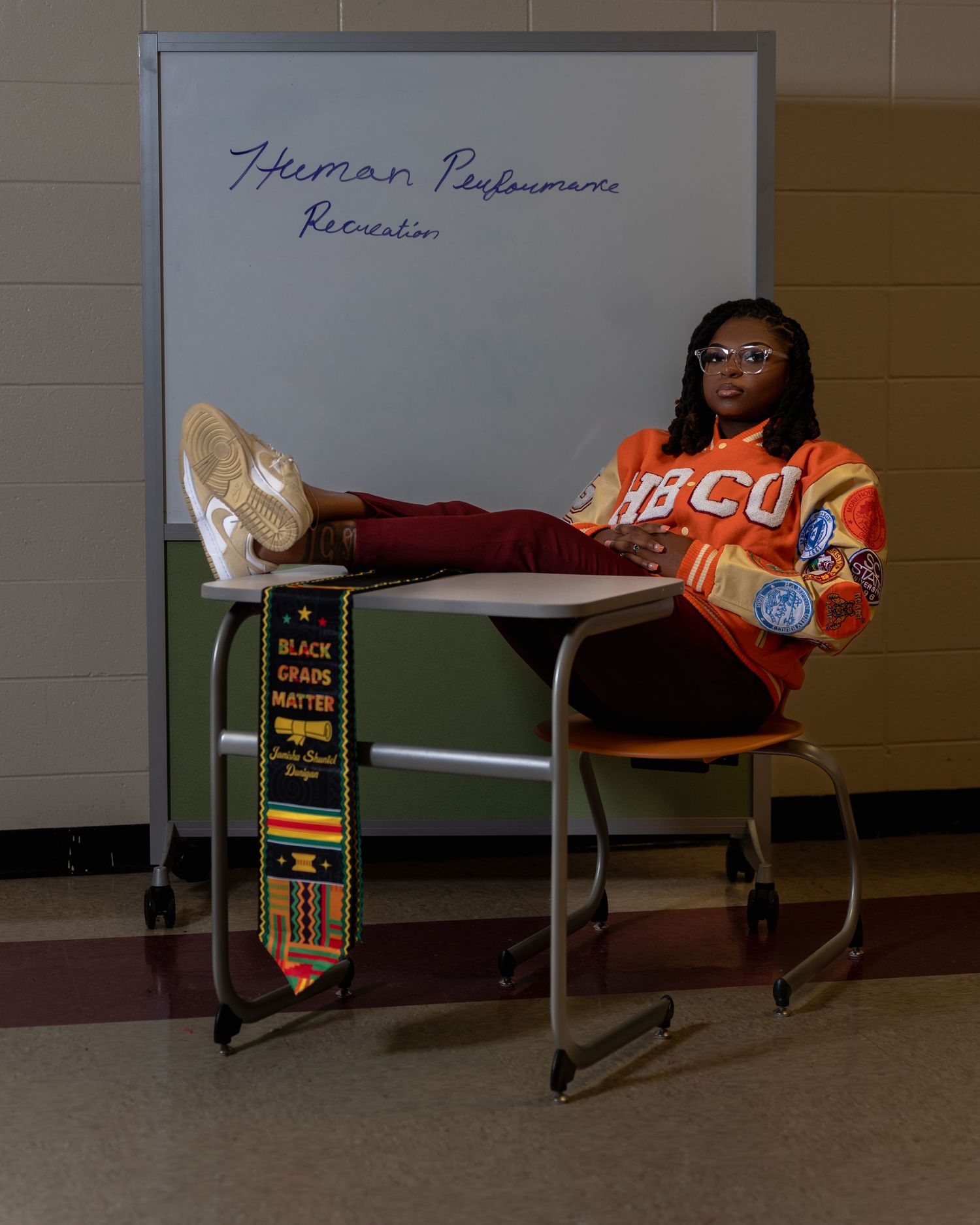 Graduate in HBCU orange jacket sitting at school desk with Black Grad Matters kente stole.