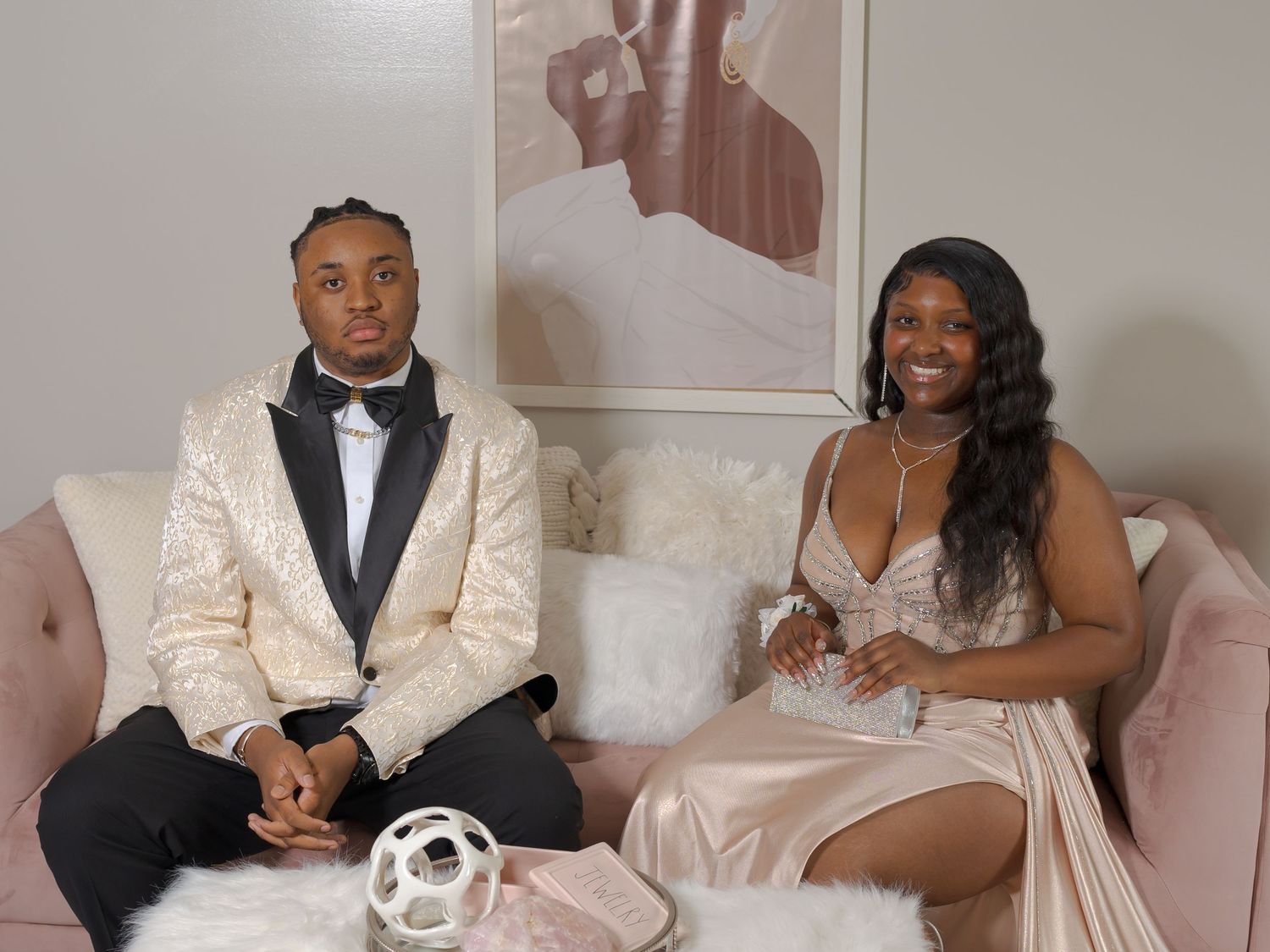 A couple dressed in formal prom attire sits together on a pink velvet sofa in a stylish indoor setting.