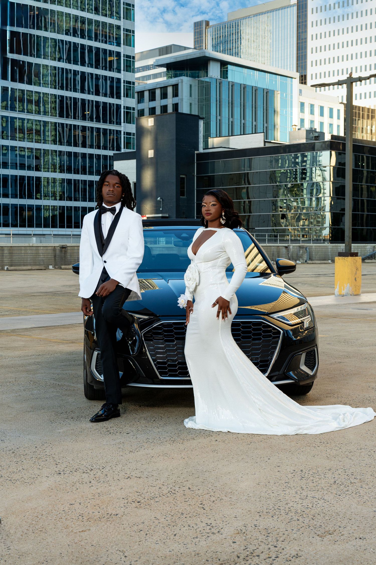 Stylish prom couple in white formal attire posing in front of black Audi with city buildings.