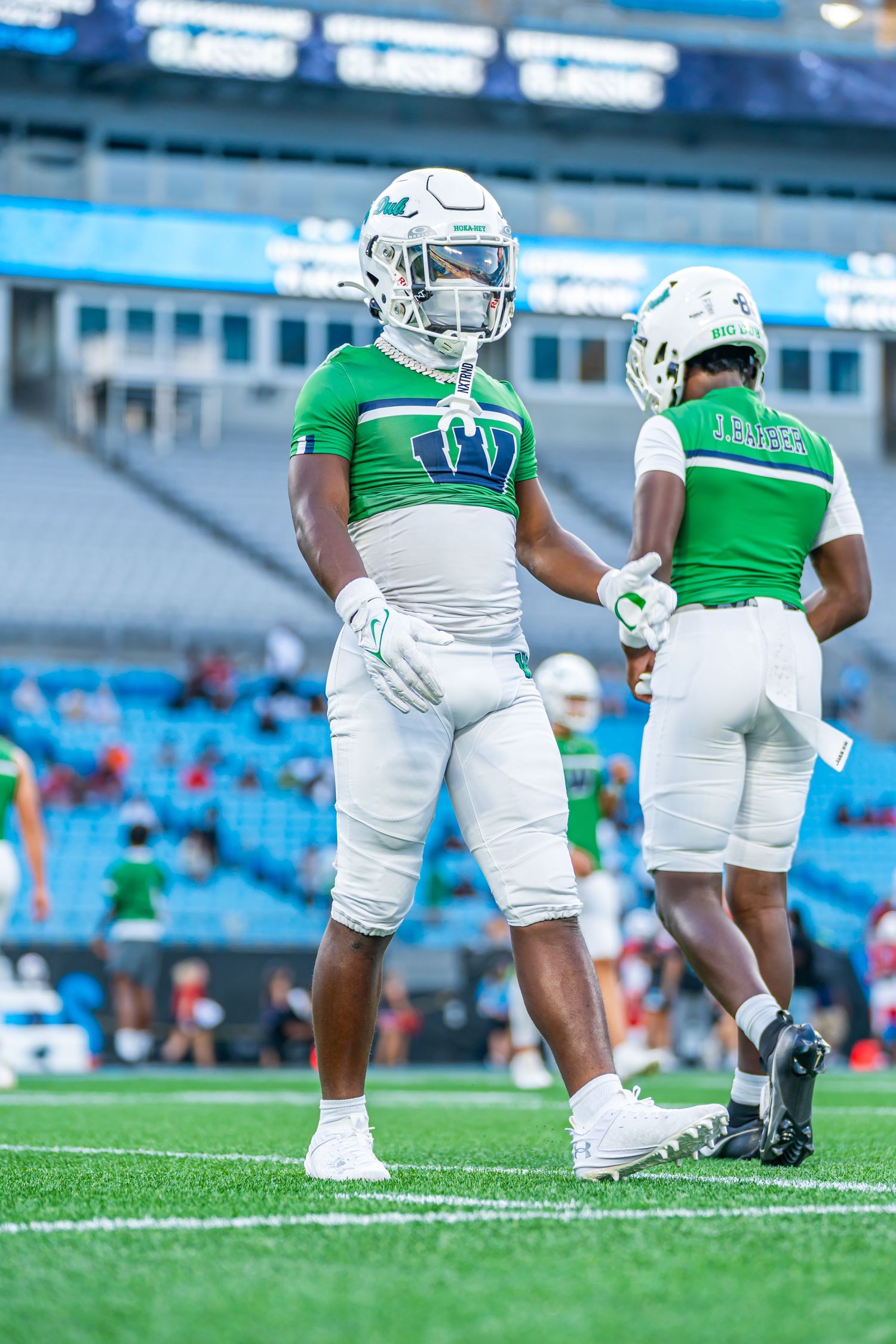 Two football players in green and white uniforms warm up on the field inside a large stadium.