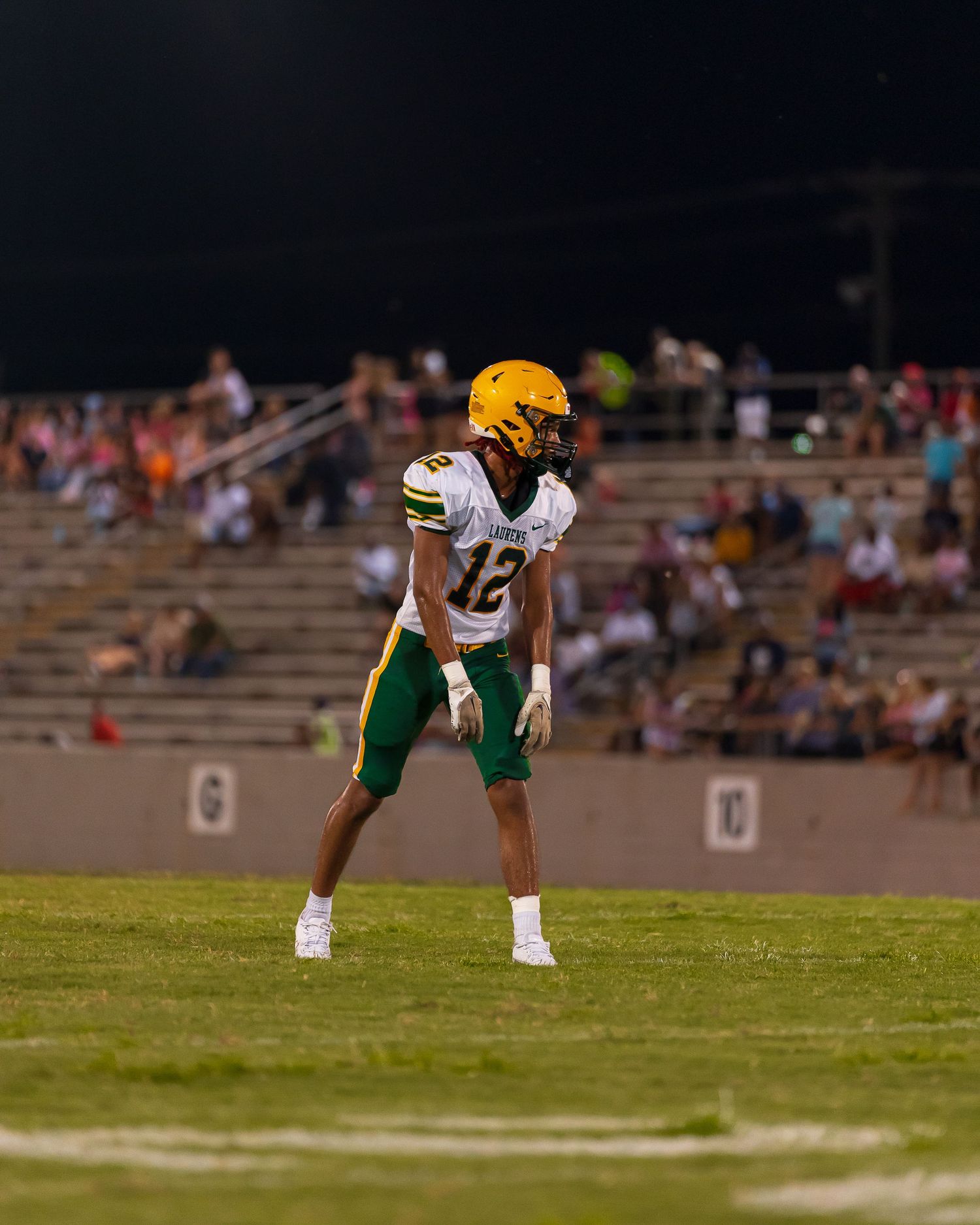 Football player wearing number 12 in green and gold uniform stands ready on field during night game.