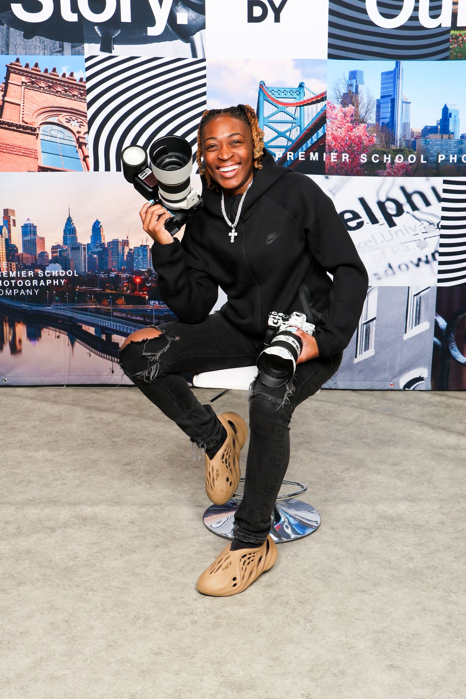Smiling young photographer poses with camera equipment at Premier School Photography backdrop.