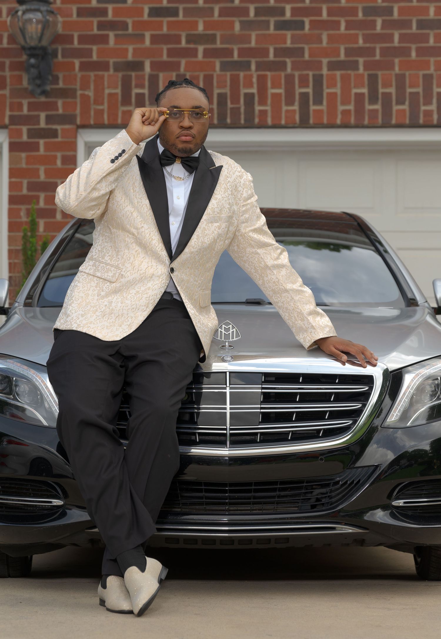 Young man in white brocade tuxedo jacket leaning on Mercedes-Maybach hood before prom night.