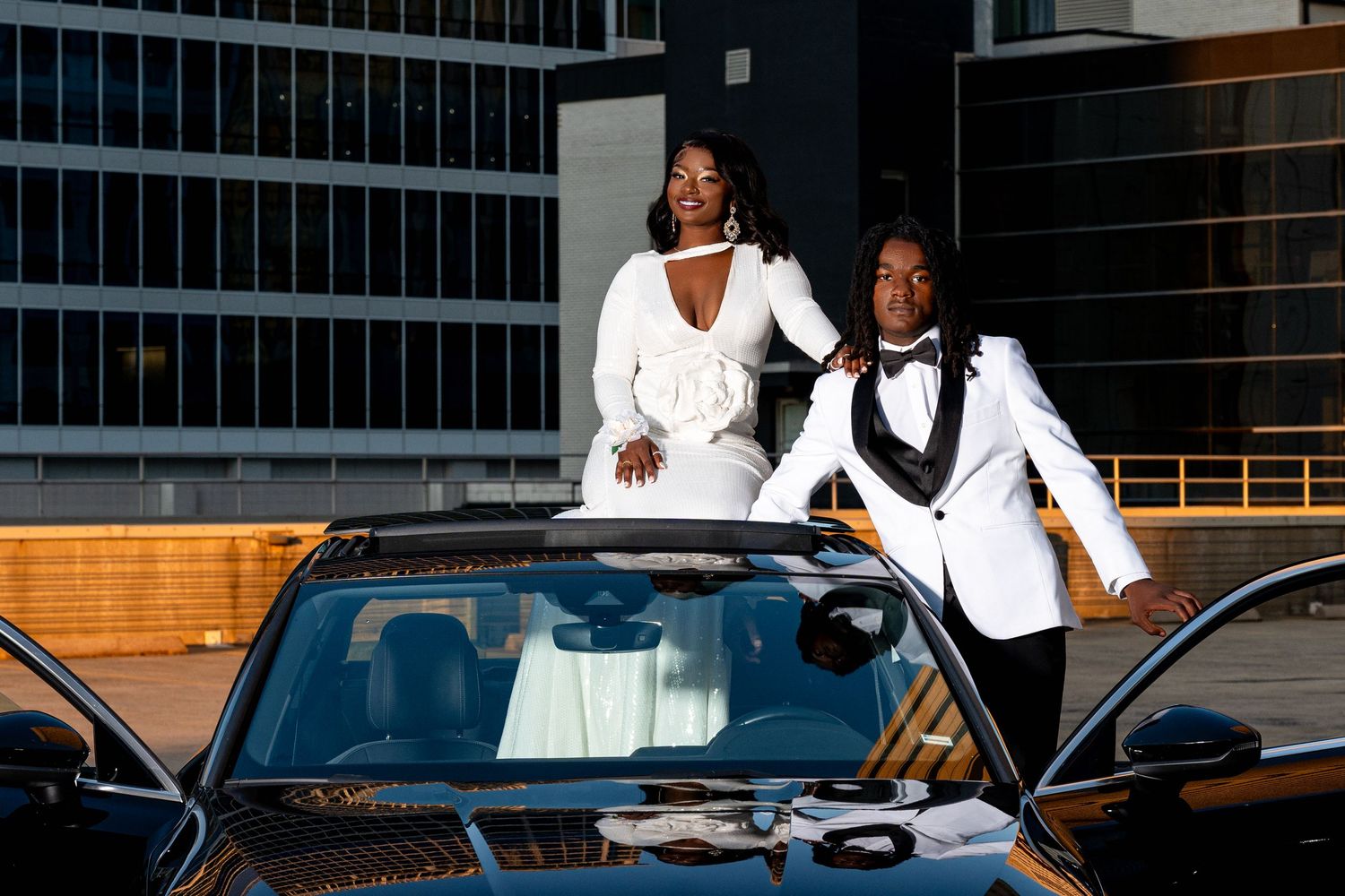 Prom couple in matching white formal outfits posing with black convertible on rooftop at golden hour.