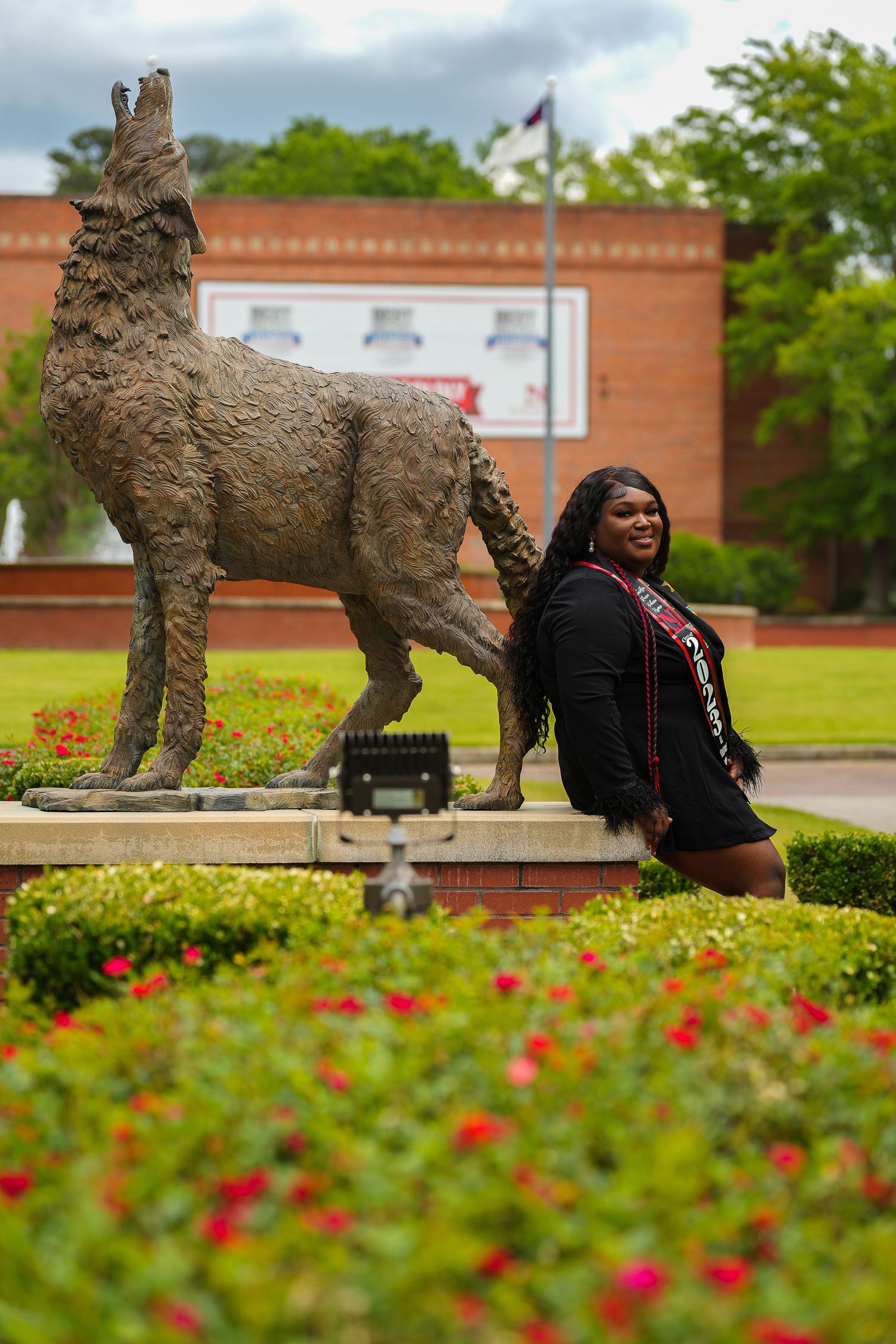 Smiling Black female graduate leaning against wolf statue on college campus surrounded by colorful flowers.