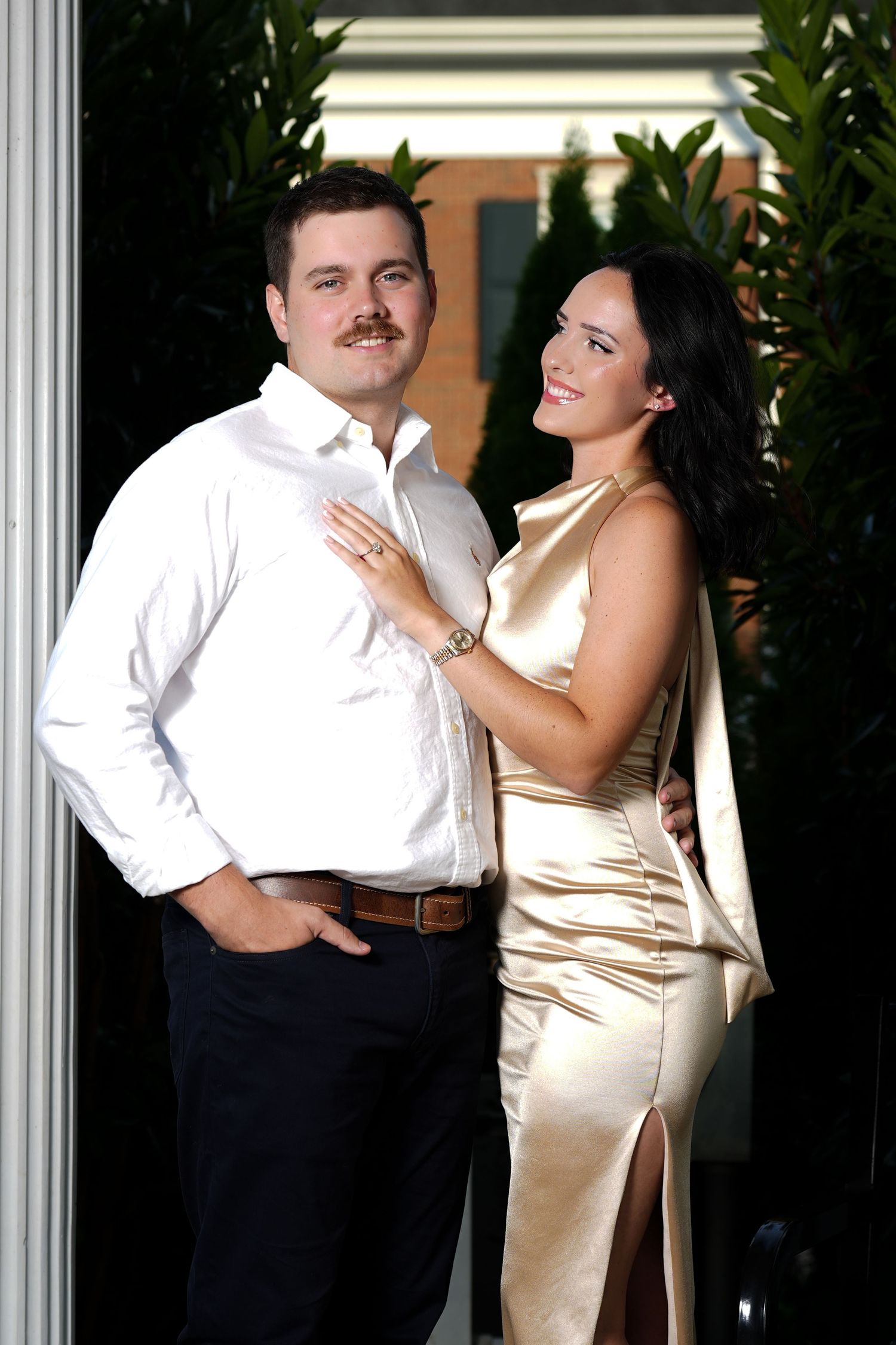 Smiling couple posing outdoors, man in white shirt and woman in gold satin gown.